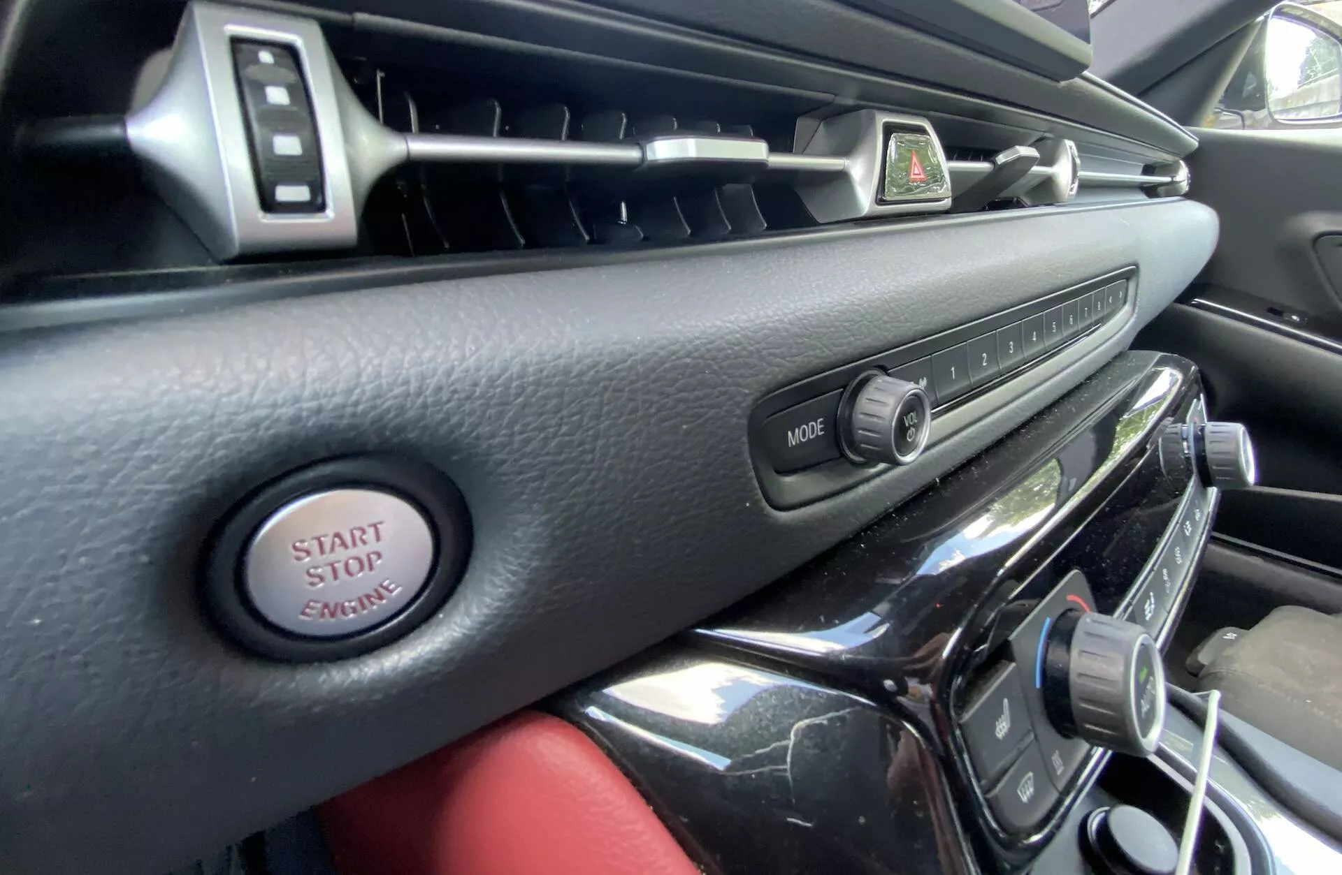 Pohled do interiéru Toyoty Supra 2019 z pohledu řidiče, zobrazující palubní desku a středovou konzoli s větráky s chromovanými akcenty, tlačítkem pro výstražné světlo, tlačítkem MODE pro multimediální systém, startovacím tlačítkem a vysoce lesklým povrchem, vyzařující luxus a modernost.