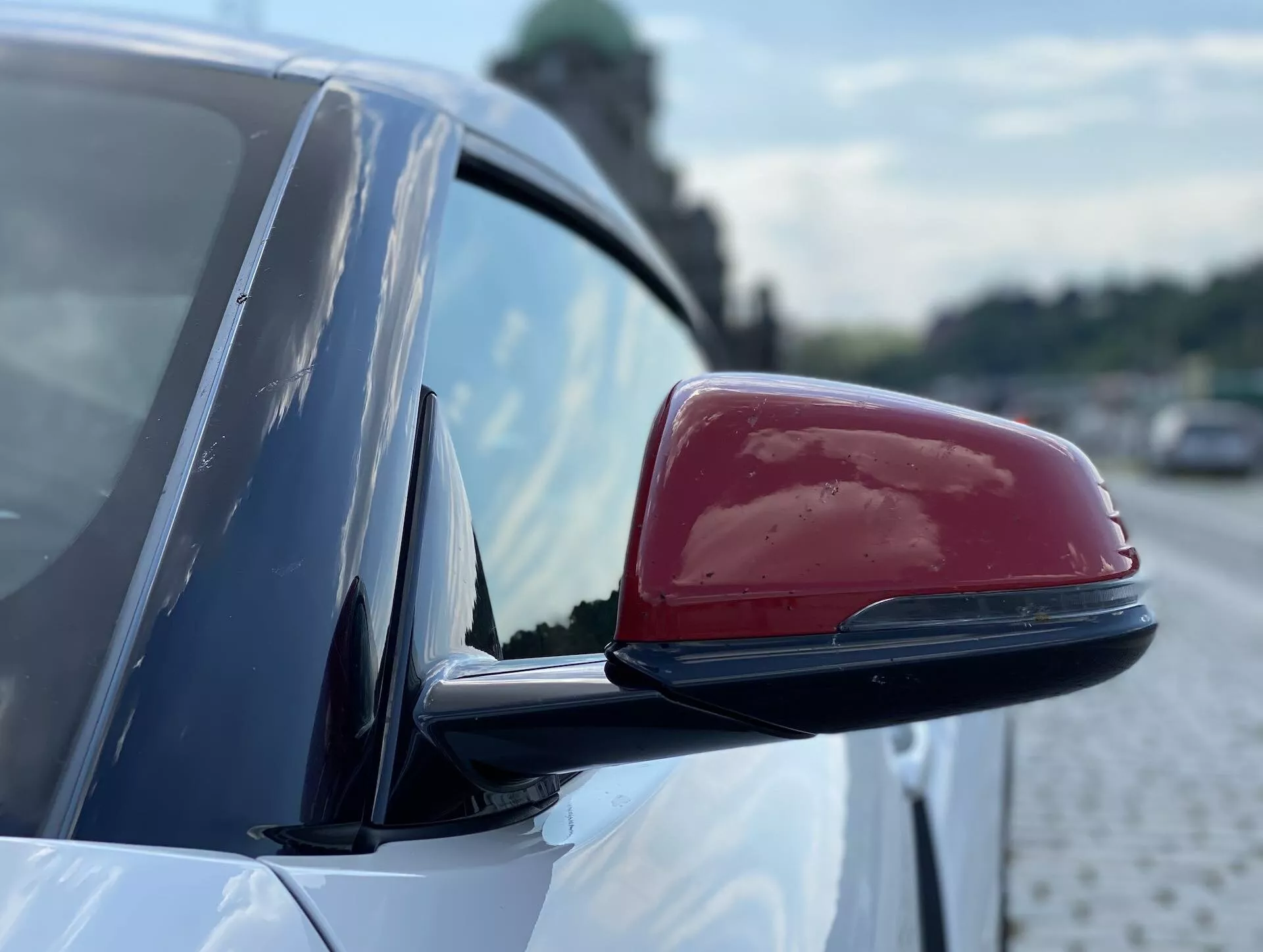 Detailní záběr na boční zrcátko vozu Toyota Supra 2019 s lesklým červeným krytem na bílém podkladu. Aerodynamický design zrcátka obsahuje možné integrované směrovky, indikátory mrtvého úhlu nebo automatickou stmívací schopnost. Rozmazané pozadí zdůrazňuje tvary zrcátka, s náznaky dlážděného povrchu a vegetace, které naznačují městské prostředí u silnice.