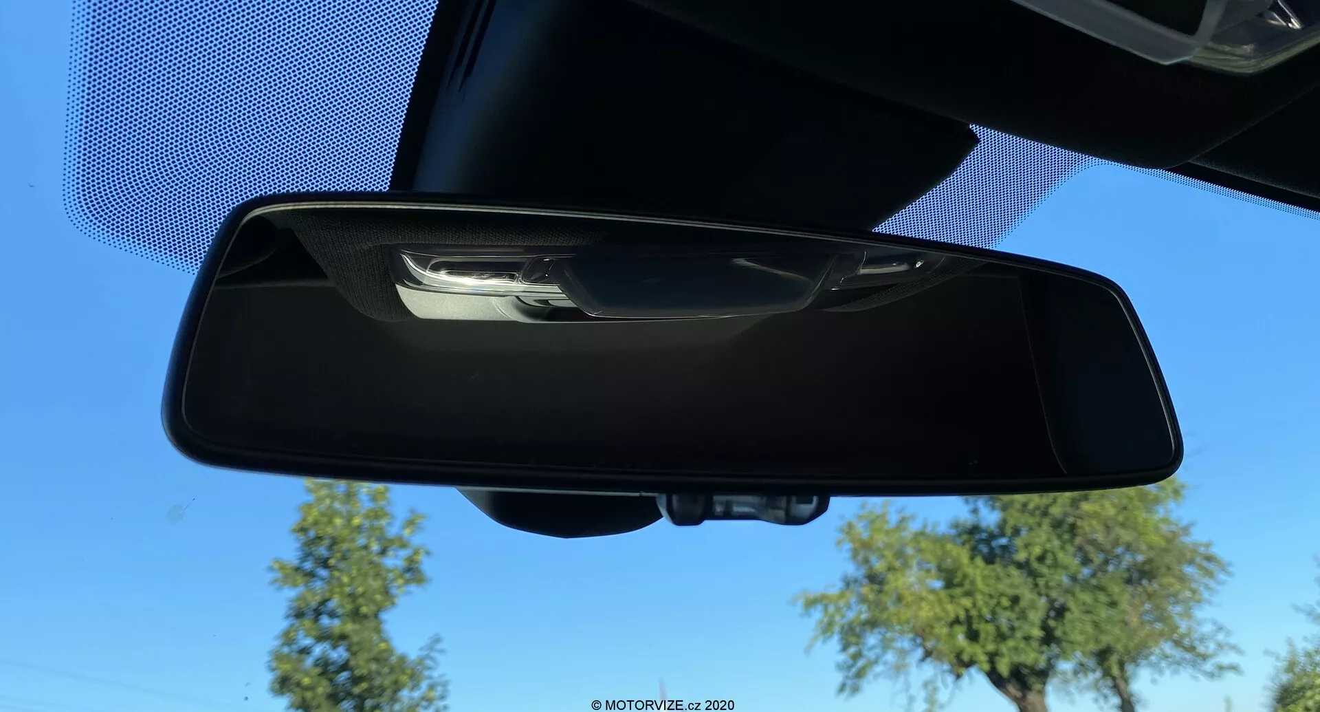Vnitřní zpětné zrcátko vozu Toyota GR Supra 2.0 Pure Automatic z roku 2019, pohled z horní přední perspektivy se zobrazením modré oblohy, stromů a části stropu vozu. Zrcátko má černý plastový rám a ruční překlápěcí mechanismus; nejsou vidět žádné pokročilé funkce jako automatické ztmavování nebo zabudované displeje.