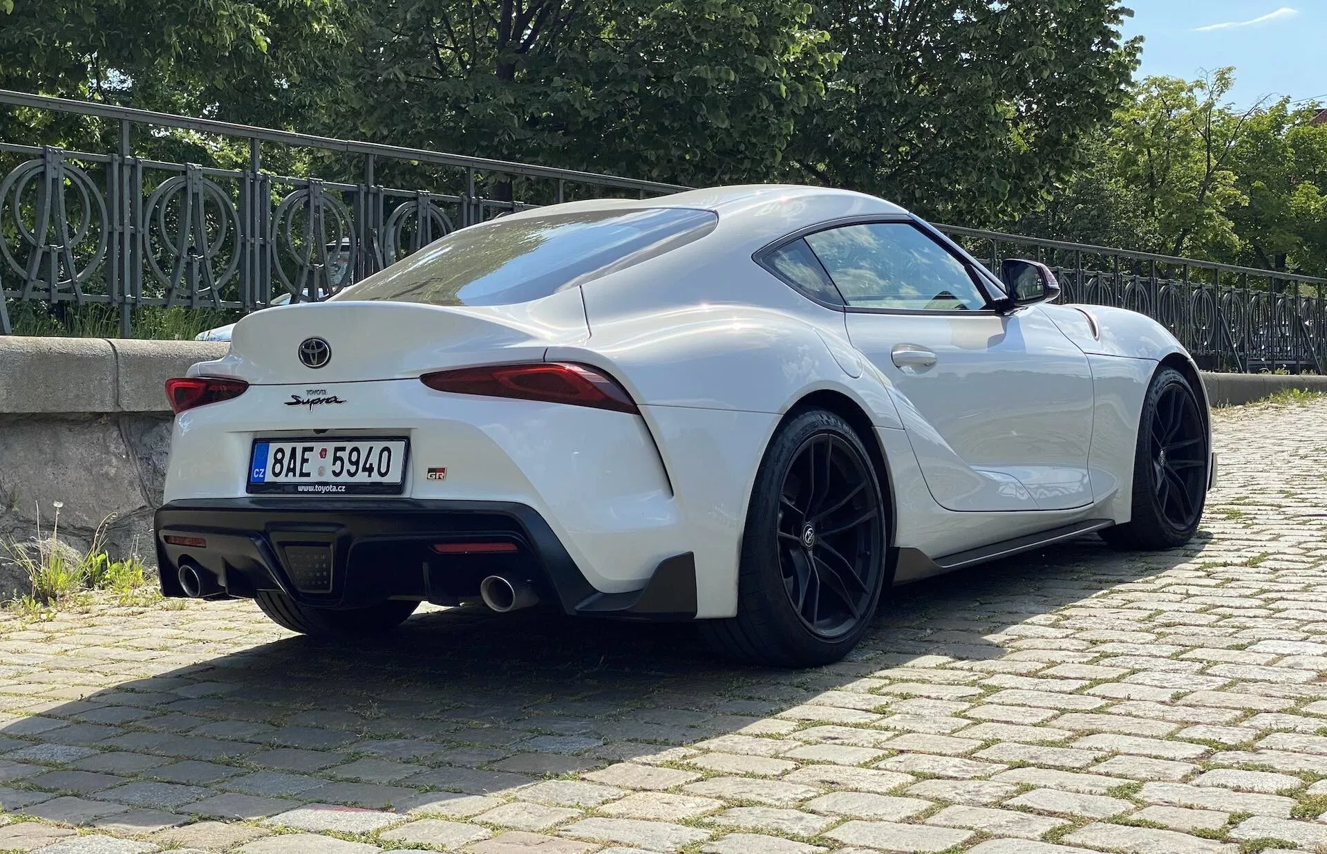 Obrázek sportovního vozu Toyota GR Supra 2.0 Pure Automatic 2019 v městském prostředí, fotografovaný z úhlu tři čtvrtin zezadu. Elegantní design zahrnuje střechu ve stylu fastback, ostře řezaná LED zadní světla, výrazný zadní spoiler, dvojitý výfuk a černá vícepaprsková kola s nízkoprofilovými pneumatikami, což naznačuje vysoký výkon a pokročilou aerodynamiku.