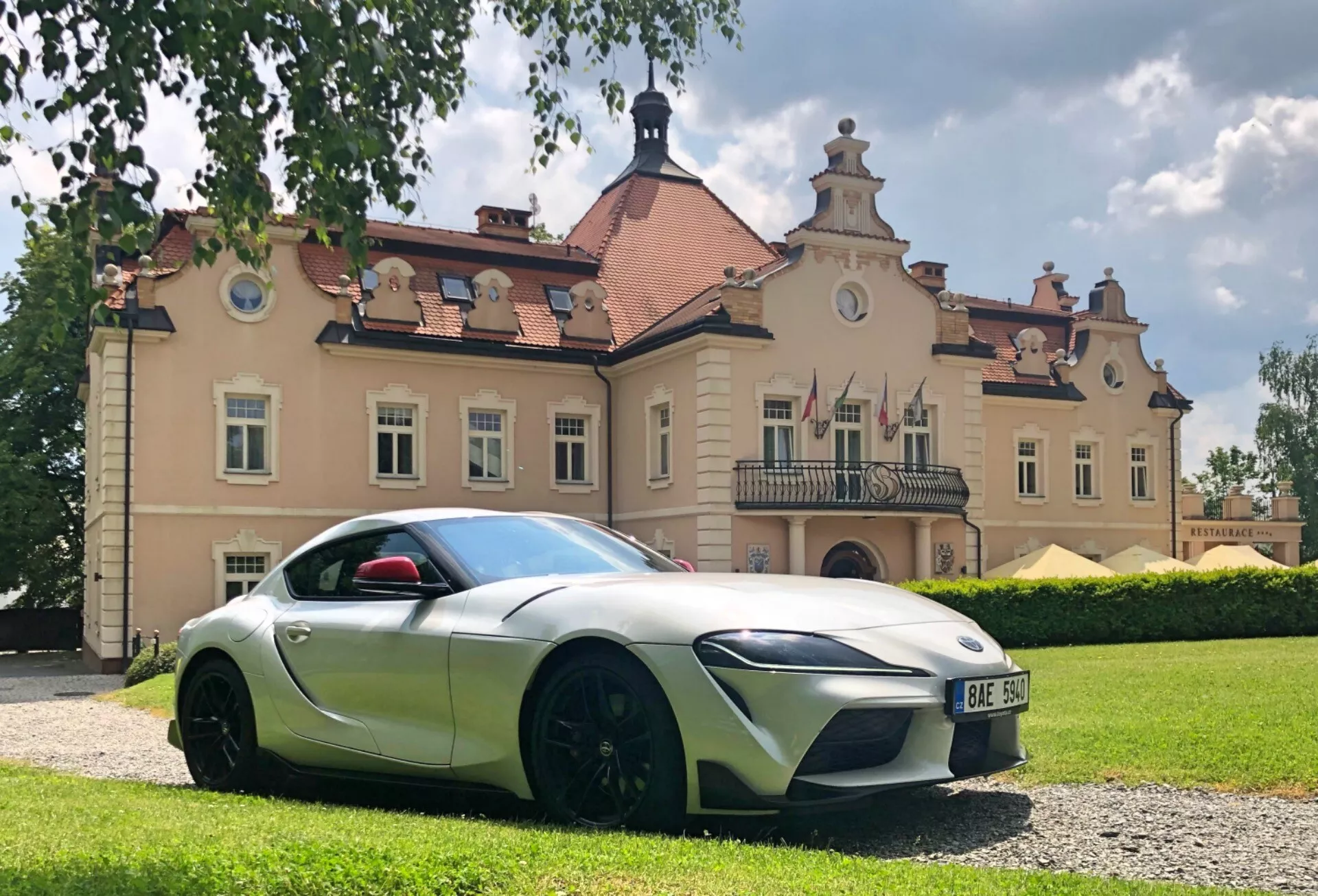 Fotografie pořízená z nízkého úhlu zachycující profil vozu Toyota GR Supra 2.0 Pure Automatic z roku 2019 z pohledu z přední strany řidiče. Elegantní, aerodynamické linie a tvarovaná karoserie naznačují sportovní design. Mezi rysy patří dvoubarevné provedení s hlavním tělem ve stříbrné/světle šedé barvě a černými akcenty, charakteristické LED světlomety, velké nasávací otvory, černá zpětná zrcátka, zapuštěné kliky dveří a velká kola s množstvím černých paprsků a nízkoprofilovými pneumatikami. Červený kryt zpětného zrcátka řidiče přidává kontrastní rozbušku barvy. Vůz stojí na štěrkovém povrchu před historickou budovou, pod částečně zataženou oblohou, což zvýrazňuje jeho křivky a designové prvky.