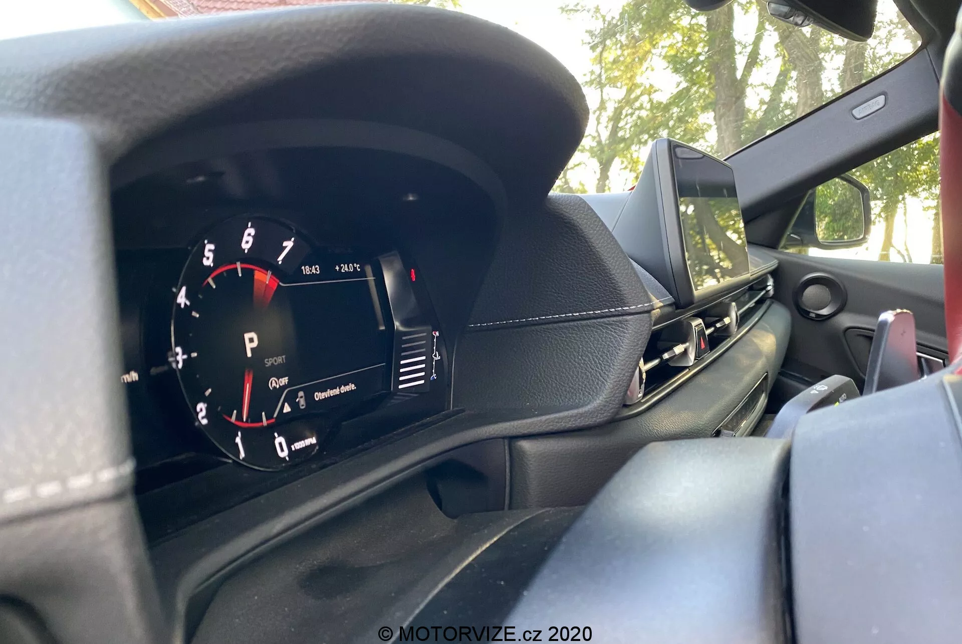 Pohled do interiéru Toyoty GR Supra 2.0 Pure Automatic z roku 2019 zaměřený na přístrojový panel za volantem, foceno z místa řidiče. Analogový otáčkoměr označený v krocích po 1 000 ot./min., s červeným polem začínajícím před 7 000 ot./min., je vidět vlevo. Digitální displej ukazuje 'P' pro parkovací režim vpravo. Palubní deska má černý texturovaný povrch, vidět je část obrazovky infotainmentu, výdech ventilace u spolujezdce a možný reproduktor na palubní desce. Část volantu, který naznačuje sportovní design, se objevuje vlevo v záběru. Venku jsou přes čelní sklo vidět stromy a obloha, což naznačuje, že vůz je zaparkován venku.