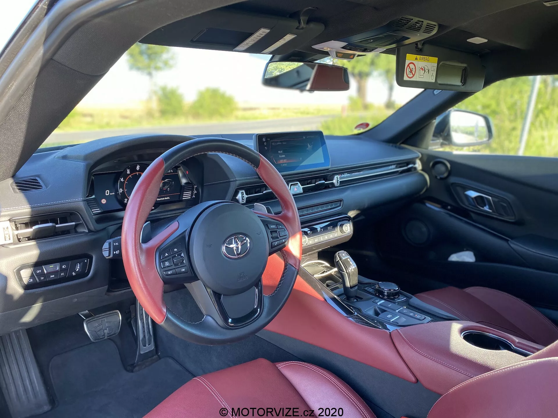 Pohled na interiér vozidla Toyota GR Supra 2.0 Pure Automatic z roku 2019, zachycující mírně necentrální pohled směrem dozadu z zadního sedadla na straně spolujezdce. Zvýrazněno červeným volantem s ovládacími prvky, digitálním přístrojovým panelem, centrálním dotykovým displejem infotainment systému, částečně viditelným ovládáním klimatizace, řadící pákou s blízkým ovládacím knoflíkem a luxusními červenými koženými sedadly, což naznačuje nadstandardní výbavu. Obrázek zdůrazňuje moderní automobilové technologie a prvky, jako je infotainment systém, digitální displej a ovládání na volantu.