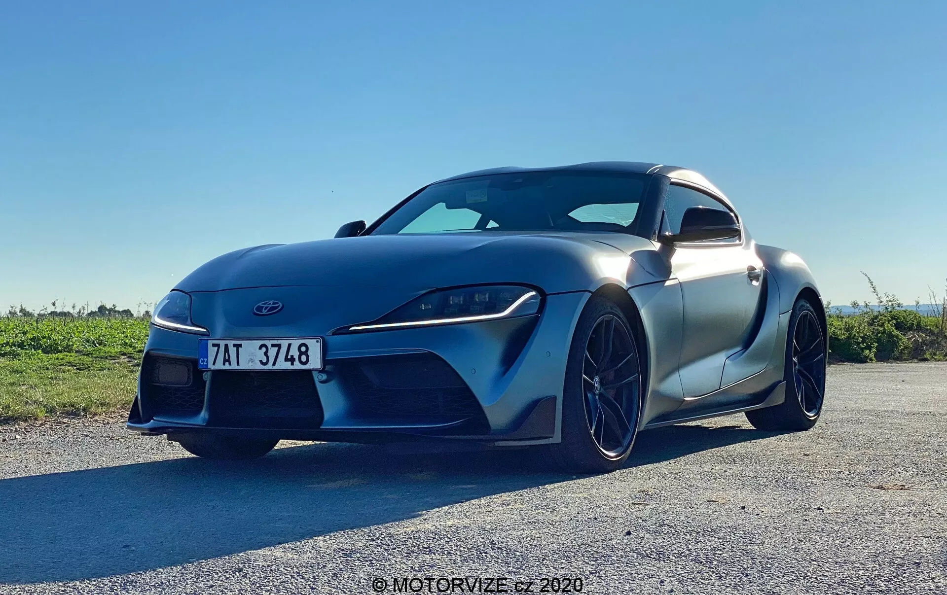 Tento obraz zobrazuje snímek sportovního vozu Toyota GR Supra 2019 2.0 Pure Automatic z nízkého úhlu, pořízený za denního světla pod jasnou oblohou. Zdůrazňuje agresivní a sportovní postoj vozu z předního levého čtvrtého úhlu, kde je vidět aerodynamický design s viditelnými prvky, jako jsou přední část s reflektory, mřížka chladiče, přední nárazník, dveře na straně řidiče, boční zrcátko, část zadního blatníku, obrysy kapoty a přední kolo se slitinou disku. Auto vyplňuje většinu rámu, což naznačuje blízký přístup, který zdůrazňuje jeho moderní design, ostrá LED světla, velkou mřížku chladiče a matný lak.