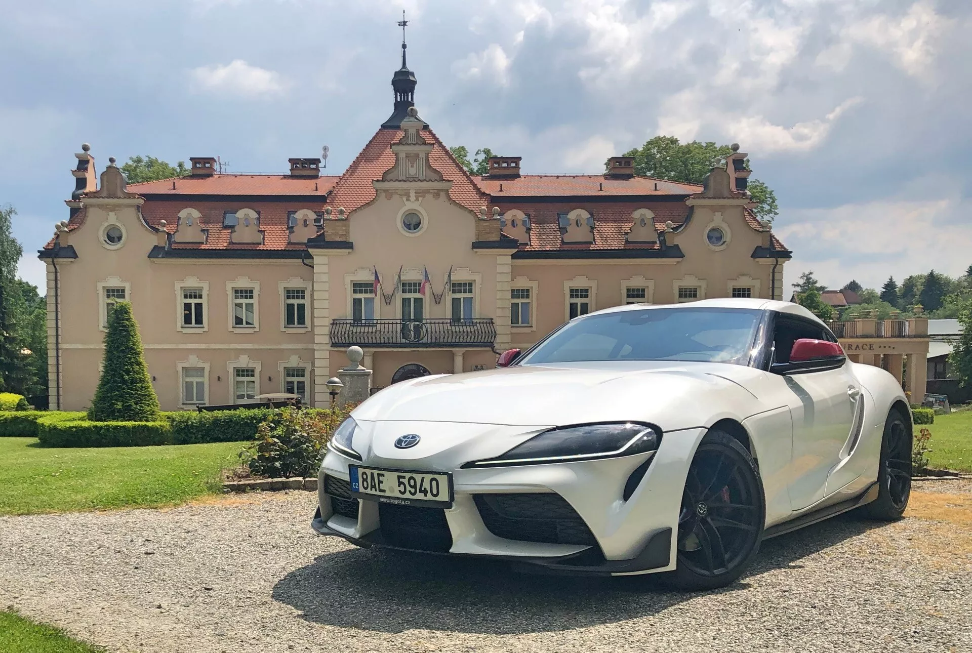 Záběr zepředu bílé sportovní vozu Toyota GR Supra 2.0 Pure Automatic 2019 se šikmými LED světlomety, výrazným předním nárazníkem s integrovanými vzduchovými vstupy, nízkou, širokou přední maskou s výrazným emblémem, velkými vícepaprskovými černými slitinovými koly, aerodynamicky navrženými zpětnými zrcátky, jemně tvarovanou kapotou a jemným zdvihem vzadu, naznačujícím spoiler. Vůz je zaparkovaný na štěrkové ploše s trávníkem v popředí, za slunného dne s ozdobnou budovou v pozadí, což naznačuje luxusní nebo historické prostředí.