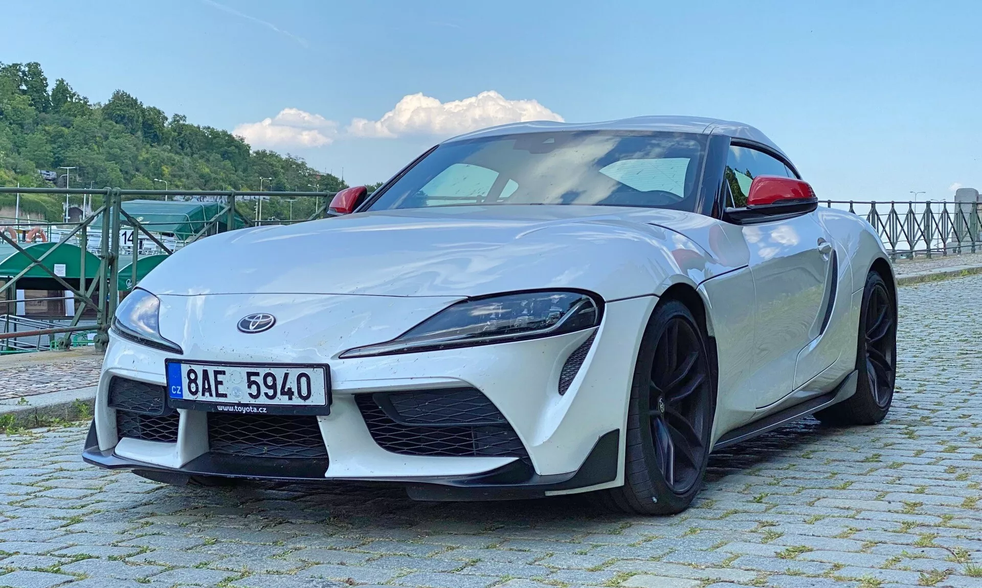 Nízký přední úhel bílého modelu Toyota Supra 2.0 Pure Automatic z roku 2019, který ukazuje klíčové vlastnosti jako jsou LED světlomety, výrazná přední mřížka, sportovní nárazník s integrovanými vzduchovody, částečně viditelné logo Supra, přední spoiler, výrazné linie kapoty, černá vícespárová litá kola odhalující vysokovýkonný brzdový systém a aerodynamická zpětná zrcátka. Střecha coupe klesá do kompaktní zadní části pod přirozeným denním světlem u vodní hladiny s kovovou strukturou v pozadí.