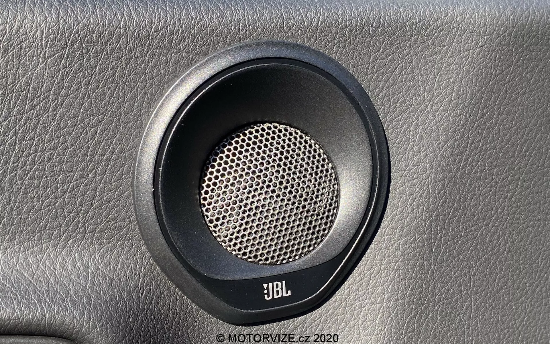 Detailní záběr reproduktoru automobilu Toyota Supra 2019 vyrobeného společností JBL, ukazující přímý pohled zepředu s mírným úhlem směrem dolů. Kulatý reproduktor má výraznou voštinovou mřížku optimalizovanou pro čistou produkci zvuku. Kolem mřížky je viditelný kruhový lem z kontrastního materiálu, pravděpodobně z gumy nebo plastu. Reproduktor je namontován na texturovaném povrchu připomínajícím kůži, což zvyšuje prémiový pocit. Logo JBL je výrazně umístěno na spodní straně, což naznačuje zaměření na vysoce kvalitní zvukové systémy.