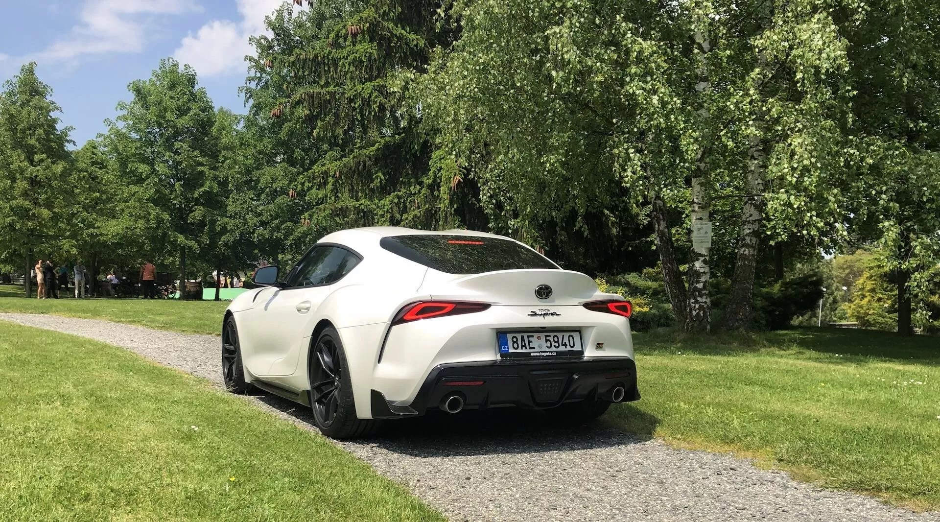 Pohled zezadu na boční stranu Toyota GR Supra 2.0 Pure Automatic z roku 2019, v bílé barvě s kontrastními černými prvky, zaparkovaný na štěrkové cestičce v parku. Obrázek zachycuje designové prvky jako zadní spoiler, dvojité koncovky výfuku, úzké LED zadní světla, sportovní litá kola a zrcátka v barvě karoserie s integrovanými směrovými světly.
