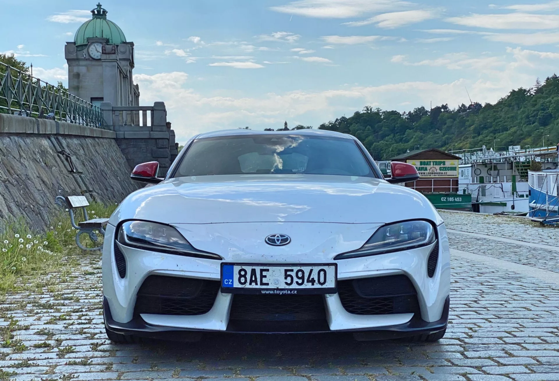 Přední záběr moderního sportovního vozu, konkrétně Toyota Supra 2019. Auto je vycentrováno v rámu a foceno z nízkého úhlu, což zvýrazňuje jeho agresivní postoj a sportovní linie. Obrázek zvýrazňuje přední nárazník, kapotu, blatníky, znak Toyoty, elegantní LED světlomety, velké nasávací otvory v nárazníku a přední splitter. Tvarovaná kapota a oblouky kol naznačují vysoce výkonný pohon zadních kol. Auto je zaparkováno na dlážděném povrchu, v venkovním prostředí s vegetací, věžovitou strukturou a architekturou u vodního okraje, pod částečně oblačnou oblohou.