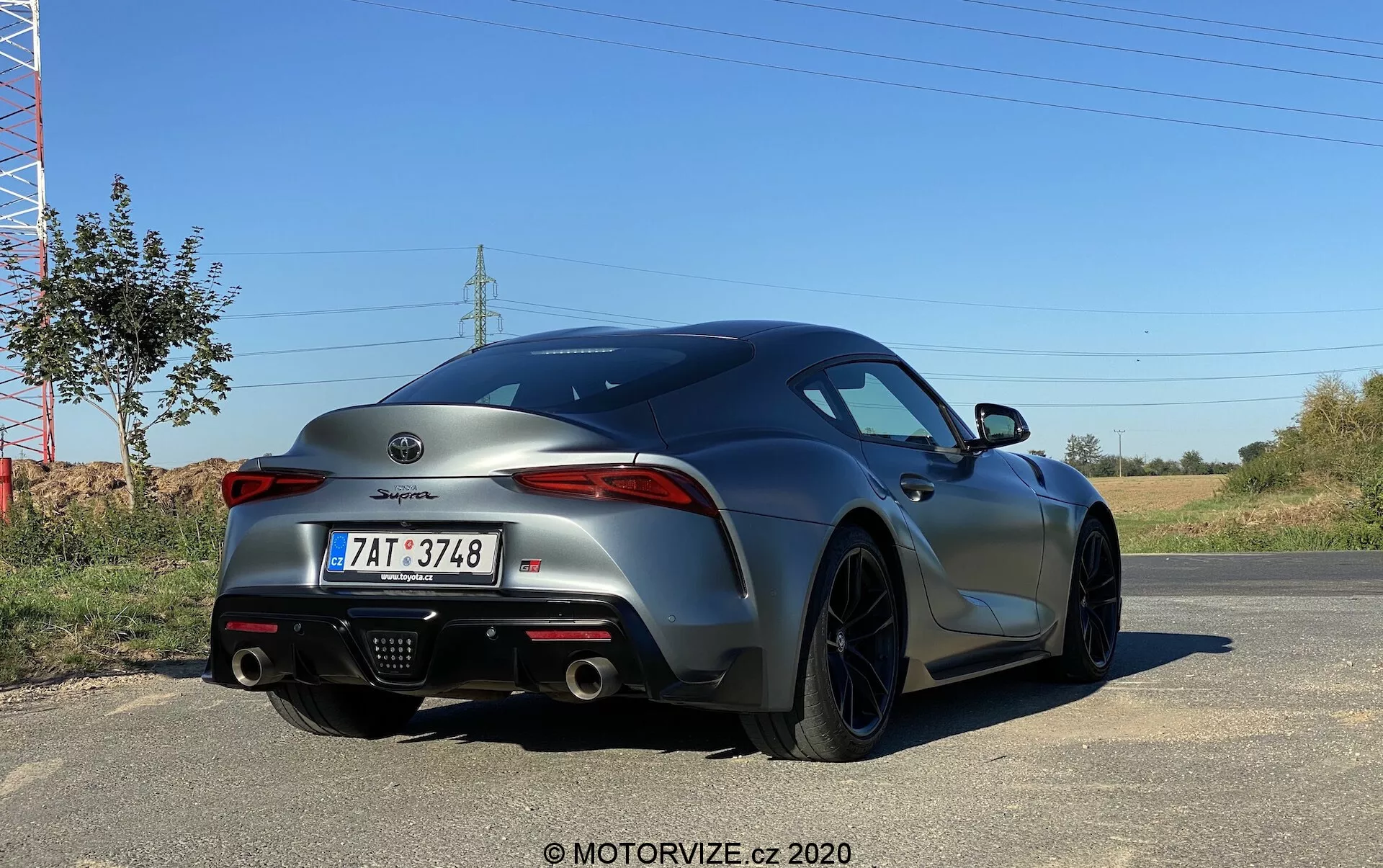 Zadní úhel sportovního vozu Toyota Supra 2019 2.0 Pure Automatic v metalické šedé barvě, ukazující jeho agresivní a sportovní zadní design, úzké směrem dovnitř namířené LED zadní světlomety, dvojitý výfukový systém uprostřed difuzoru, tmavá litá kola, evropskou poznávací značku a označení modelu na kufru.