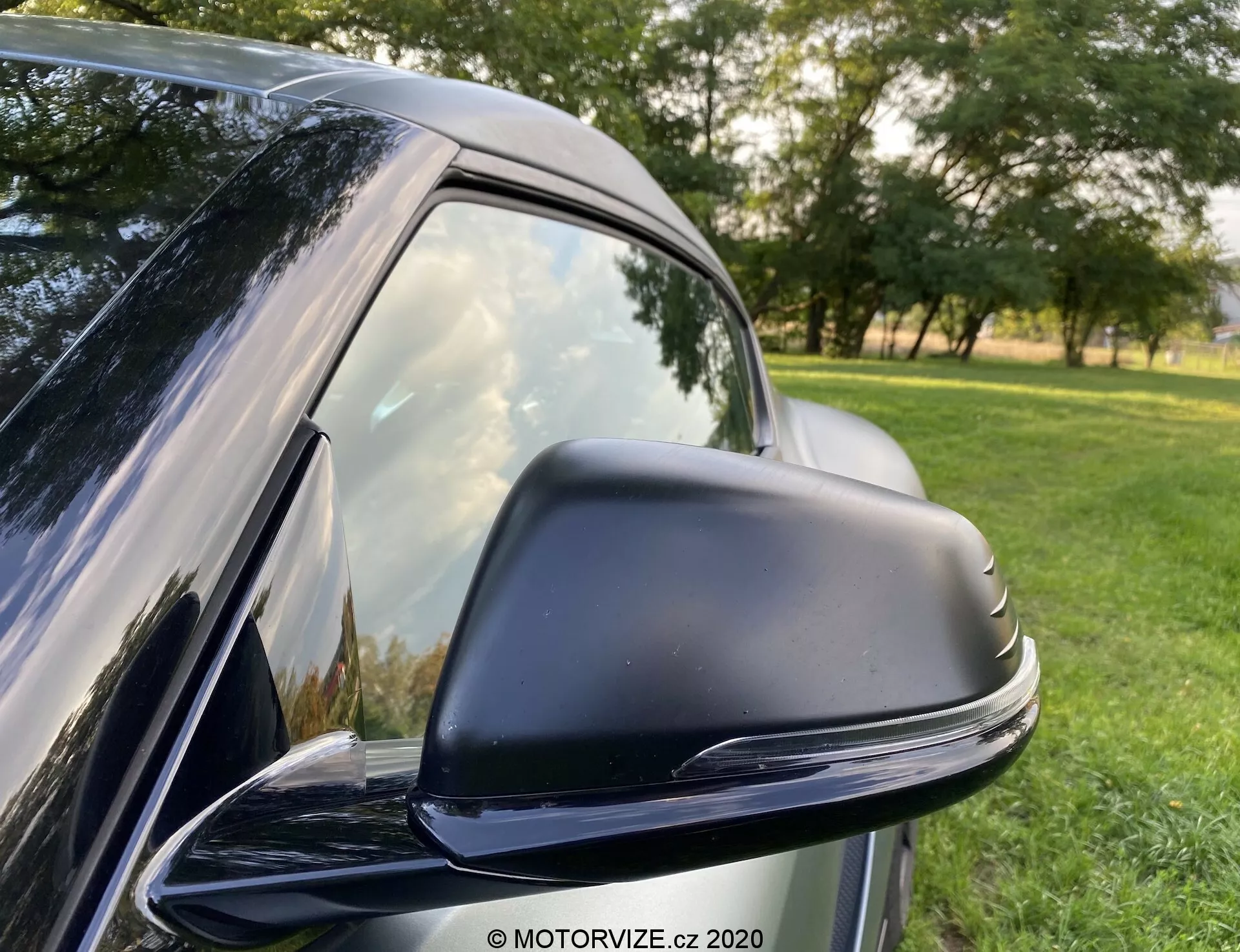 Detailní pohled na elegantní černé boční zrcátko na vozidle Toyota GR Supra 2.0 Pure Automatic model 2019, které ukazuje jeho moderní, aerodynamický design. Zrcátko je umístěno v černém krytu s tvarem optimalizovaným pro snížení odporu vzduchu a je pravděpodobně elektricky nastavitelné. Viditelná je zatmavené boční okno vozu a hladce přecházející B-sloupek. V pozadí je přírodní prostředí s trávou a stromy. Odrazy naznačují jasné venkovní podmínky.