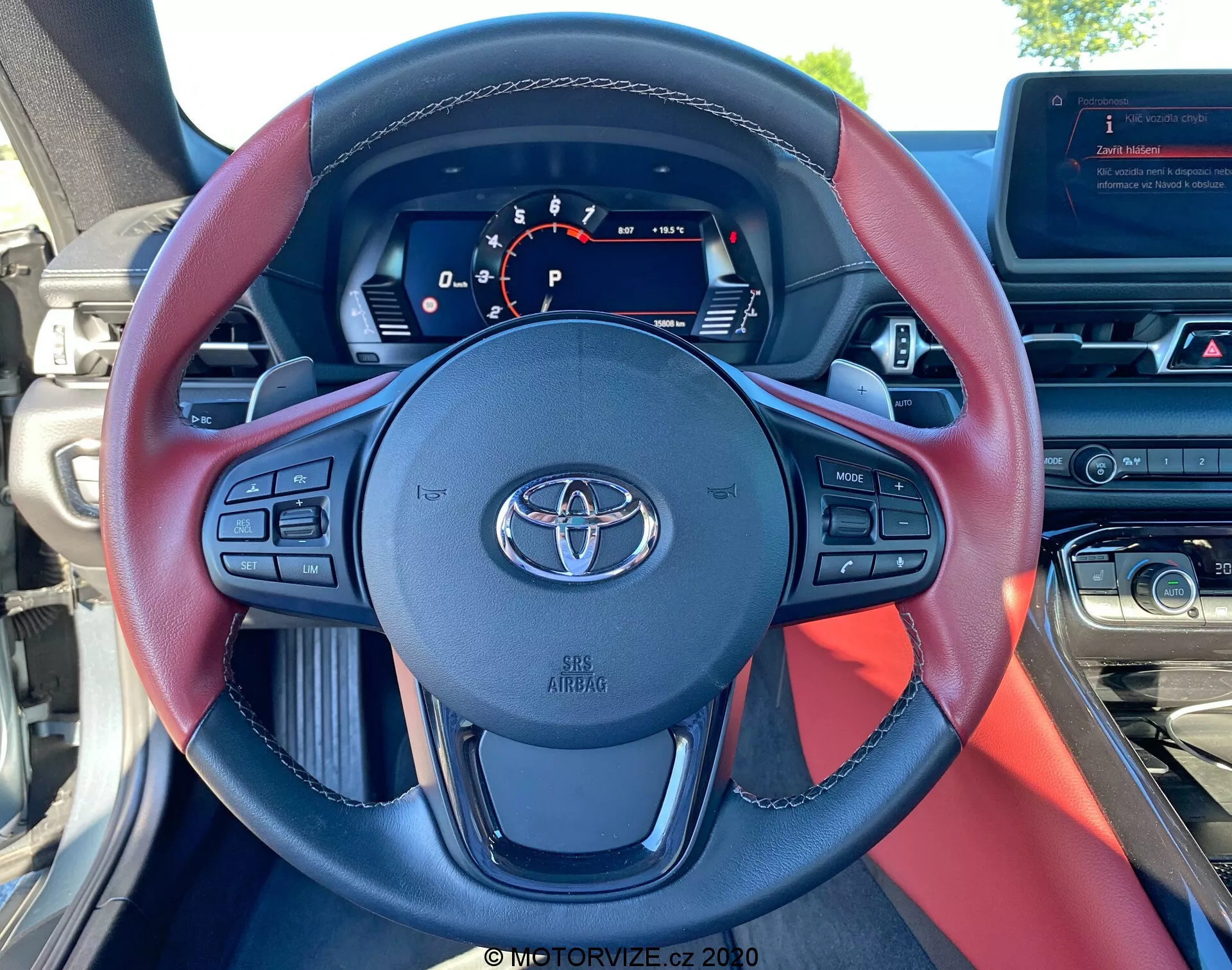 Pohled do interiéru od místa řidiče vozu Toyota Supra GR 2019 2.0 Pure Automatic, ukazující tříramenný volant s černými a červenými akcenty, znakem Toyota a různými ovládacími tlačítky pro hlasitost, hlasové ovládání, telefonní operace a adaptivní tempomat. Digitální přístrojový panel zobrazuje velký centrální rychloměr, ukazatel převodovky s 'P' a další indikátory stavu, s ukazatelem paliva viditelným vpravo nahoře. Je také viditelná část středové konzoly a informačního zobrazovacího systému, stejně jako horní část palubní desky a denní světlo skrz čelní sklo.