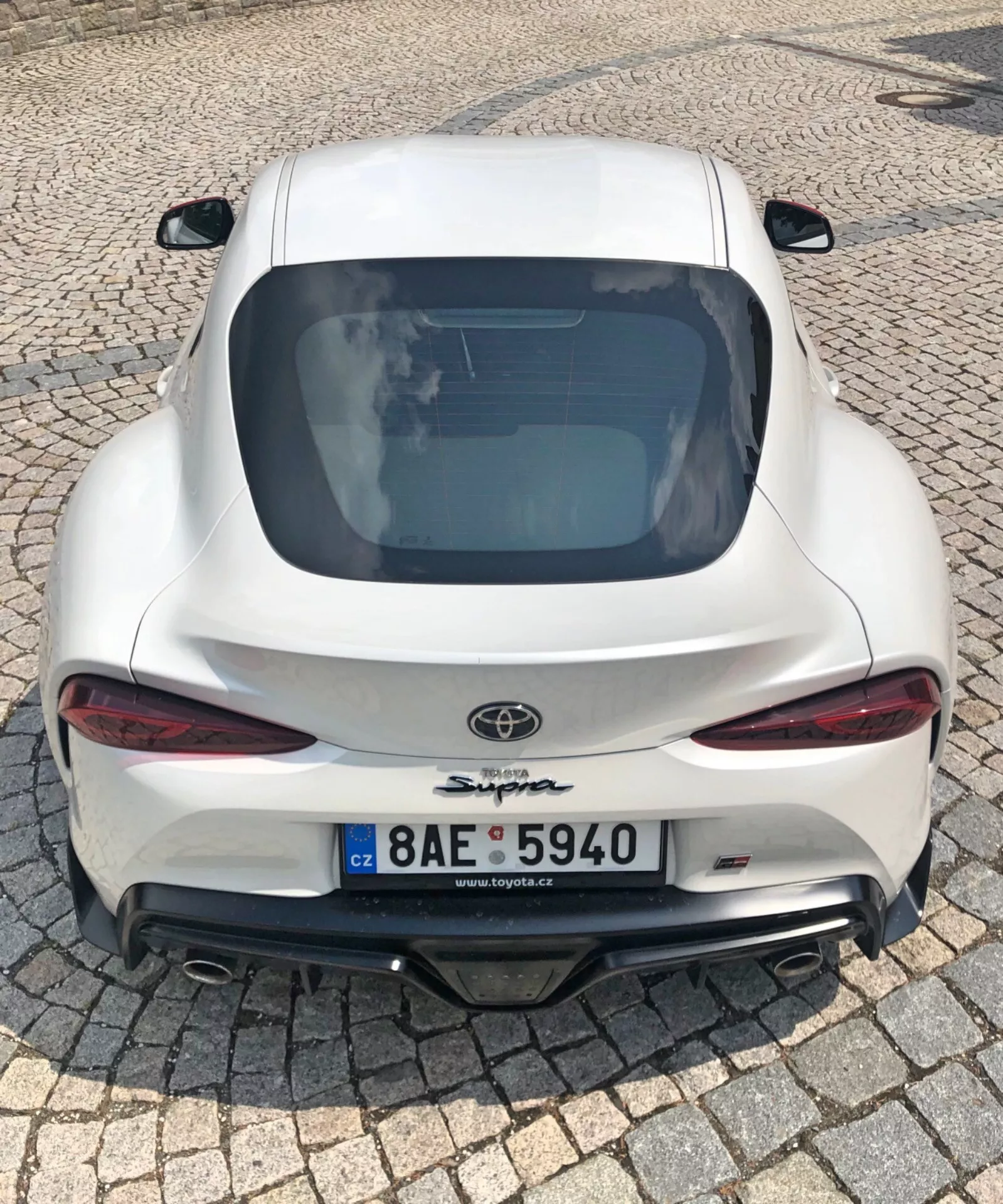Tento obrázek ukazuje zadní pohled na bílý sportovní vůz Toyota Supra 2019 v detailním záběru z vysokého úhlu, který zachycuje dvojité sportovní výfuky, aerodynamický design s integrovaným spoilerem, elegantní LED zadní světla, velký zadní difuzor, sklonenou střechu, prominentní znak Toyota a název 'Supra' a poznávací značku České republiky. Lesklý lak a moderní technologické prvky jako LED světla a aerodynamický balíček naznačují výkonný a stylový vůz v městském prostředí.