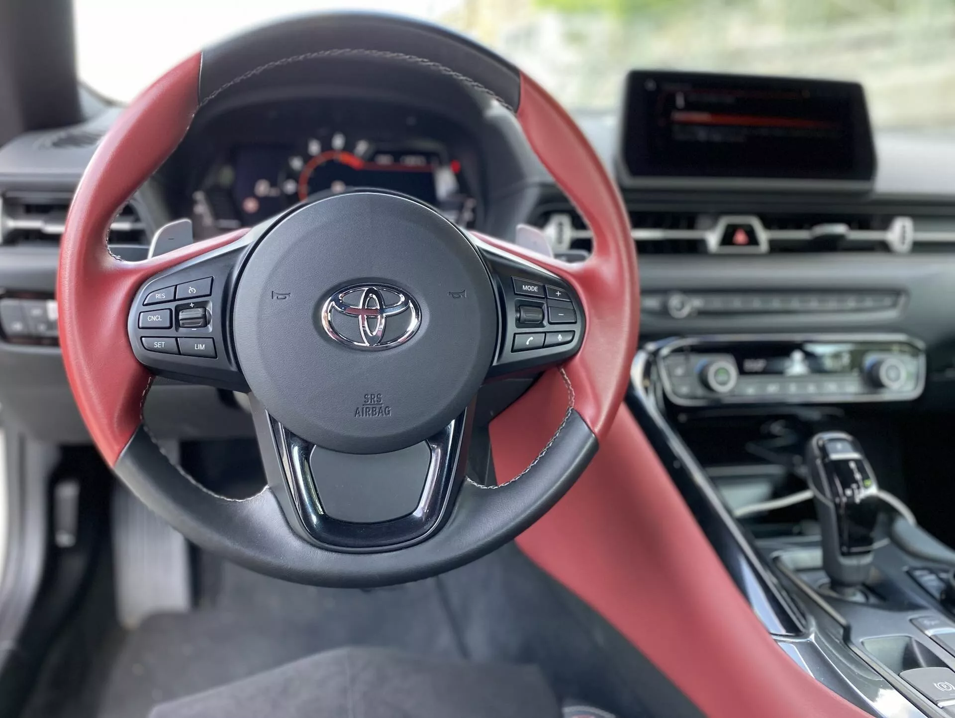 Interiérový pohled na Toyotu Supra 2.0 Pure Automatic z roku 2019. Moderní, tříramenný volant s logem Toyota a červeným a černým barevným schématem dominuje obrazu. Sofistikovaný volant nabízí ovládání audiosystému, tempomatu a hands-free komunikace. Za volantem je částečně viditelný digitální přístrojový panel, který naznačuje elegantní, technologicky pokrokový design. Středová konzole obsahuje informační a zábavní systém a tlačítka pro ovládání klimatizace. Náznak červené a černé kožené sedačky řidiče a ergonomické řadicí páky naznačuje automatickou převodovku. Denní světlo osvětluje stojící vůz, čímž zdůrazňuje jeho sportovní a luxusní interiér.