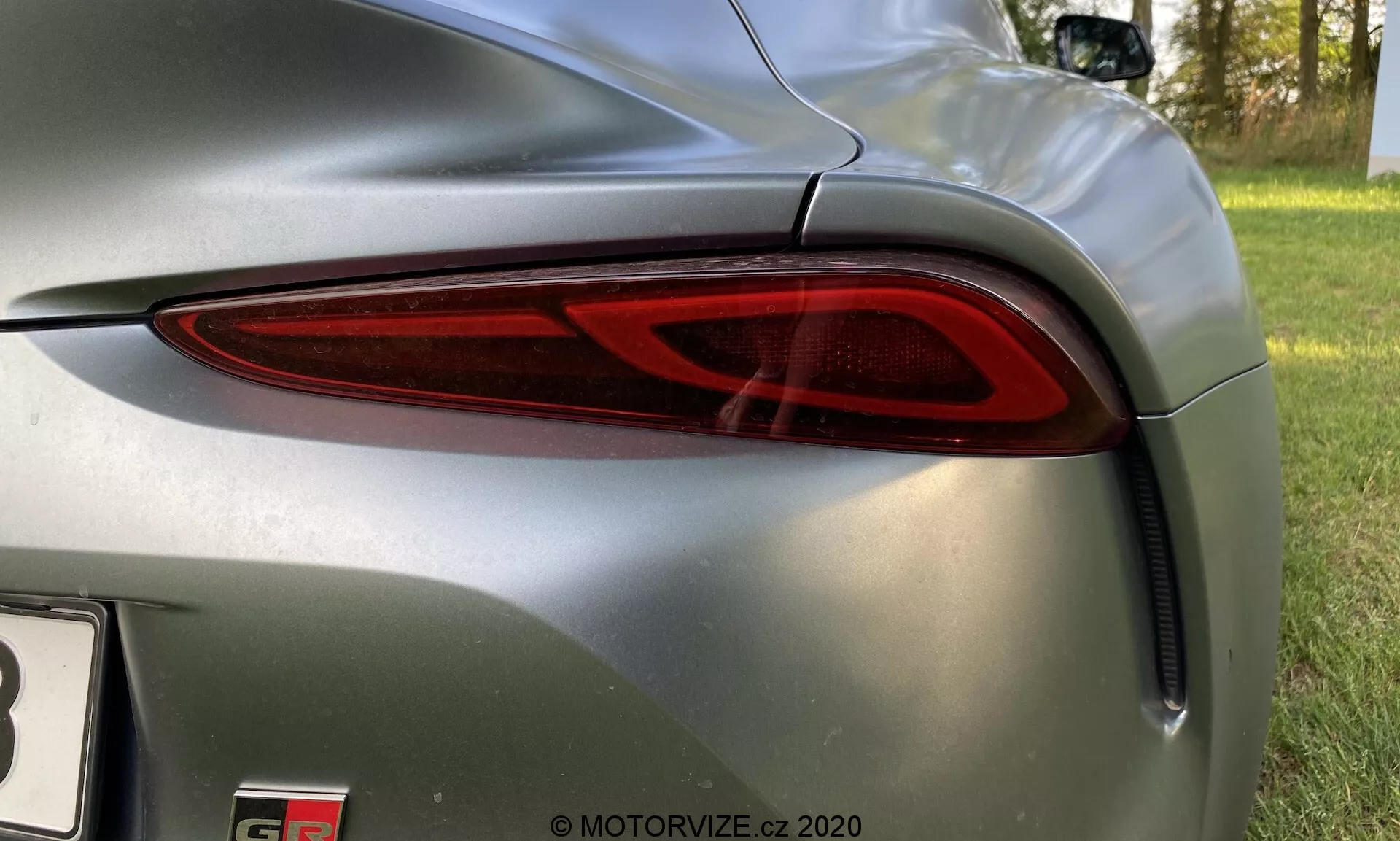 Detailní pohled na zadní levou stranu vozu Toyota GR Supra 2019, kde je moderní, elegantní zadní světlo s integrovanou LED technologií. Červený průsvitný kryt se stáčí kolem boku vozu, zlepšuje viditelnost. Kolem světla je karoserie v metalické šedé barvě s lesklým povrchem, odrážejícím okolí. Zadní nárazník pod světlem má větrací otvor, přispívající k aerodynamickému designu. Částečně viditelný modelový odznak naznačuje sportovní nebo luxusní vůz. Záběr je z mírného nadhledu, zaměřený na designové prvky vozu.