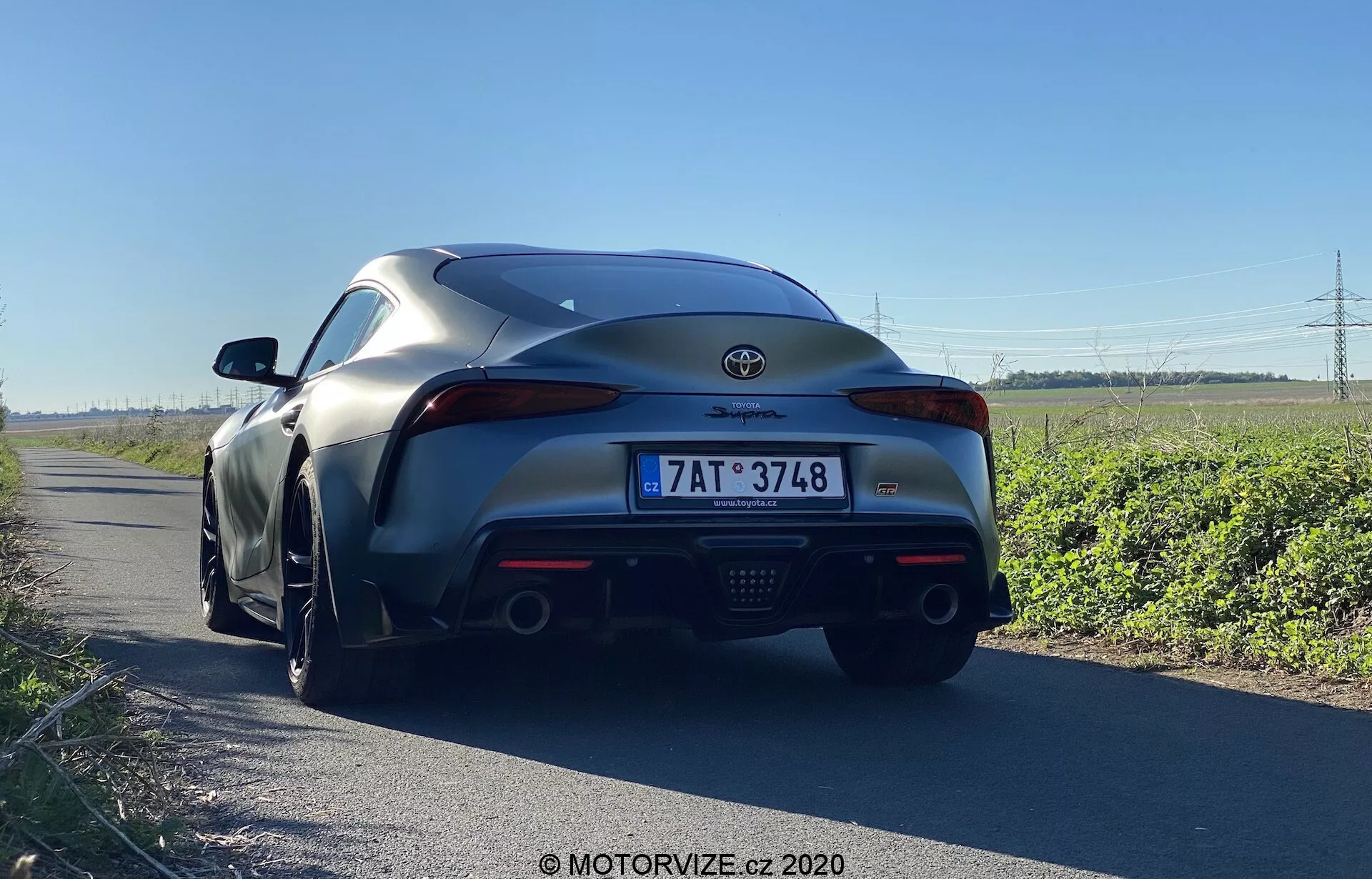 Pohled zezadu na Toyota Supra 2019, foceno z nízkého úhlu těsně nad zemí, ukazuje zadní design vozu. Detailní pohled na zadní nárazník, zadní světla, emblém výrobce, duální výfuky a zadní difuzor. Venkovní prostředí na krajnici zdůrazňující výkon a moderní designové prvky, jako jsou LED zadní světla, aerodynamický zadní spoiler a integrovaný difuzor.