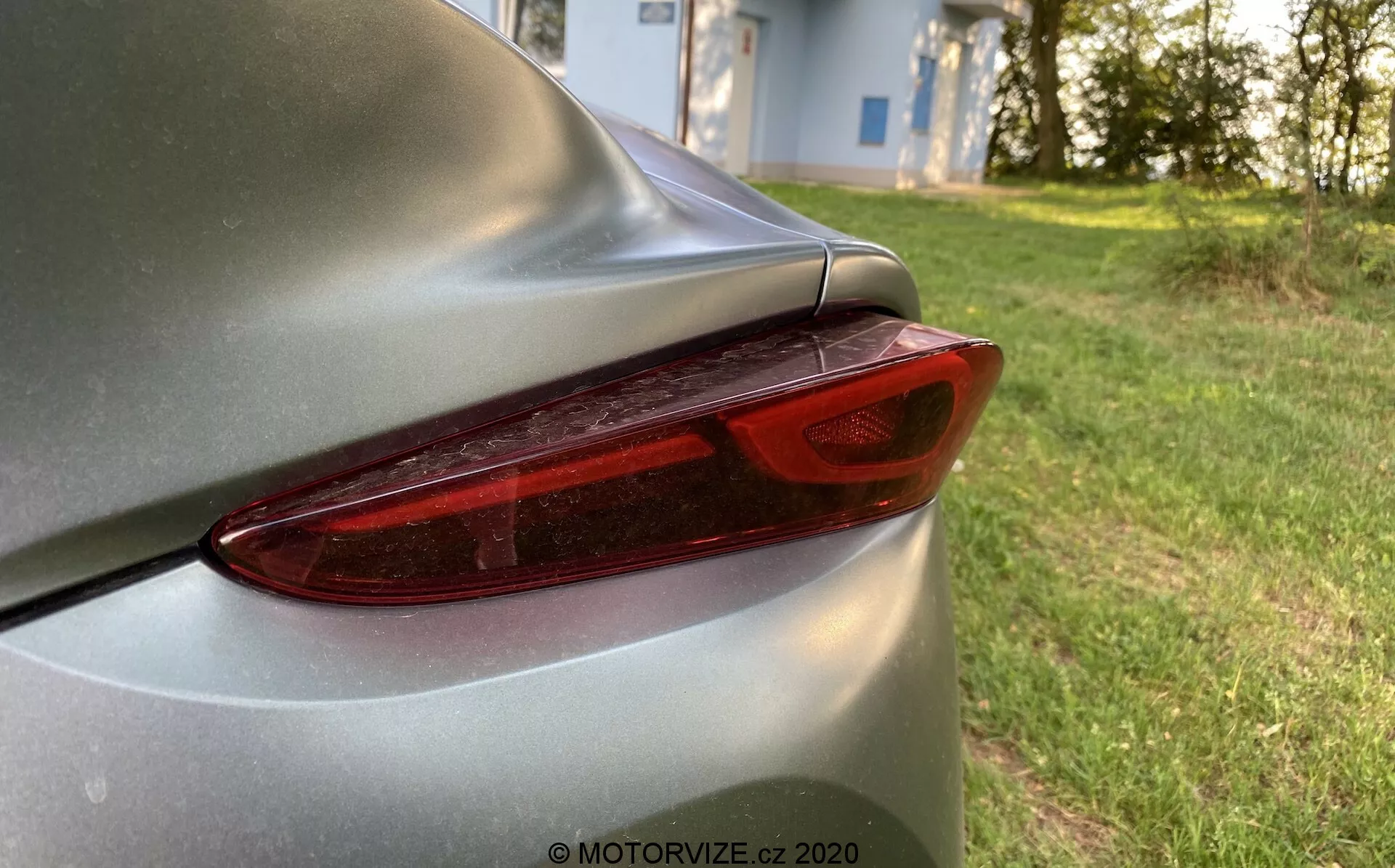 Detailní záběr zadní části vozu Toyota Supra 2019 (Toyota-GR-Supra-2-0-Pure-Automatic), zobrazující pravé zadní světlo a část víka kufru. Moderní LED design zadního světla se vyznačuje jasnými, segmentovanými liniemi pod červeným krytem a je doplněn stříbrným nebo světle šedým tělem s lesklým lakem. Aerodynamický obrys a mírný spoiler ve tvaru zdvihu okraje víka kufru zdůrazňují elegantní design vozu. Fotografie zdůrazňuje detail zadního světla s bokeh pozadím, které naznačuje přirozené, klidné venkovní prostředí, pravděpodobně poblíž obytné oblasti.