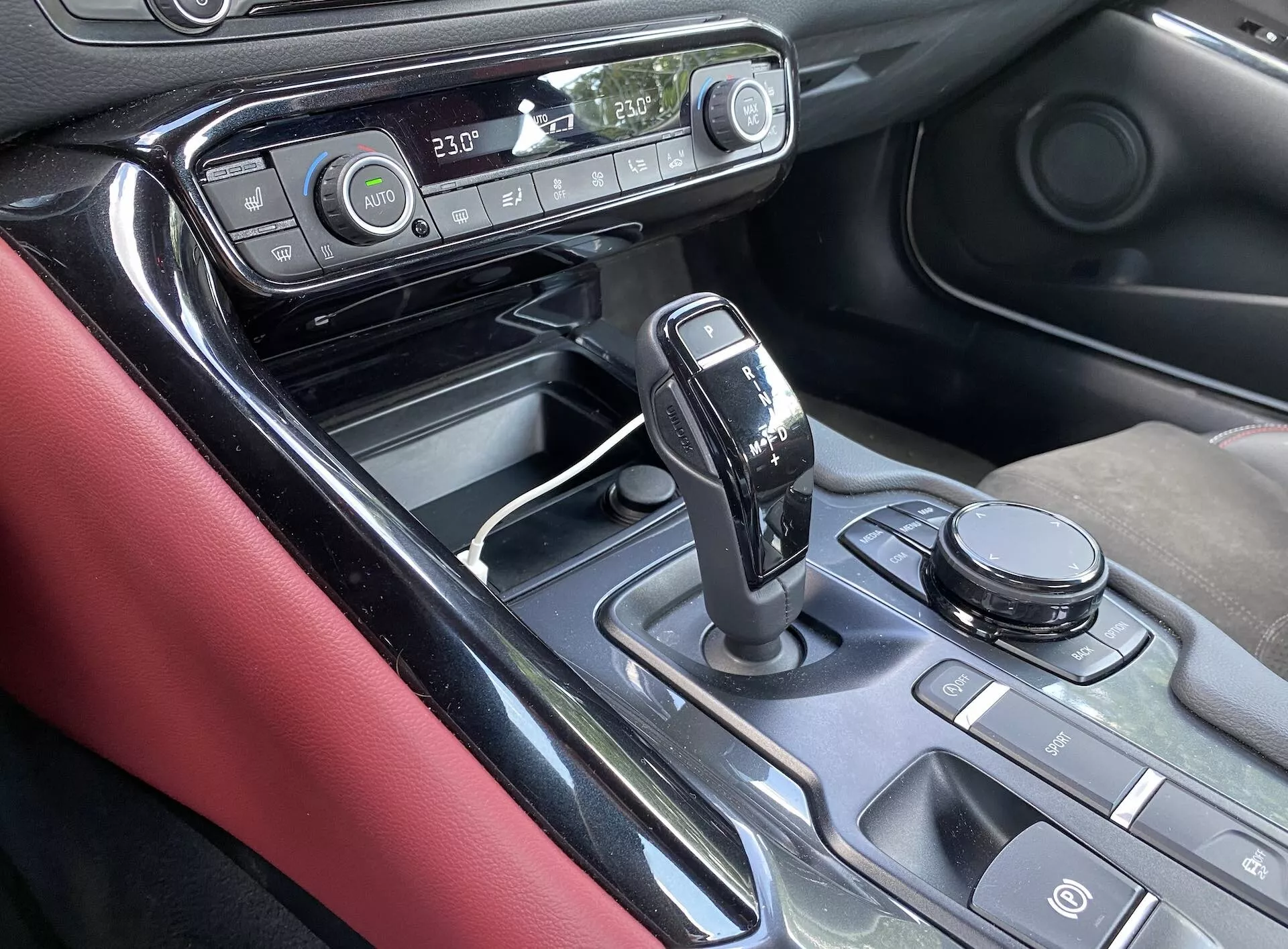 Detailní, šikmý pohled na středovou konzoli a část palubní desky Toyoty Supra z roku 2019, zaměřený na oblast řadicí páky. Pohled z místa spolujezdce zvýrazňuje moderní řadicí páku automatické převodovky s lesklým černým povrchem a chromovými detaily v poloze 'park'. Středová konzola má kovové obložení, různé tlačítka, volič jízdních režimů nebo ovládací číselník infotaiment systému, displej zobrazující nastavení teploty klimatizace, hodiny a materiály jako leskle černé, kovové akcenty a matné černé povrchy. Červené kožené obložení na vnitřní straně dveří dodává kontrast interiéru. Obrázek naznačuje pokročilý infotaiment a ovládání klimatu, ergonomické umístění ovládacích prvků a prvky luxusního designu.
