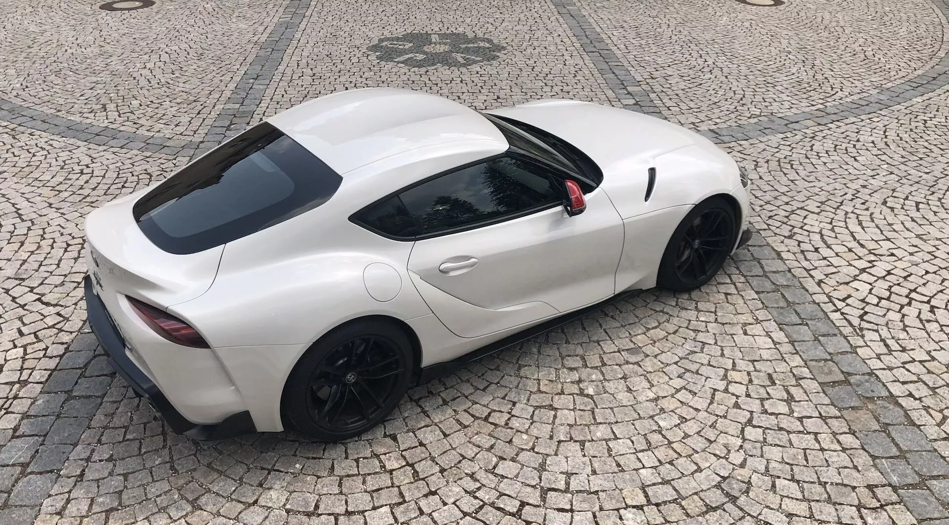 Pohled shora na elegantní bílý sportovní vůz Toyota Supra GR 2.0 Pure Automatic z roku 2019 s aerodynamickým designem, dvoudveřovou karoserií kupé a černými hliníkovými koly. Černá zpětná zrcátka a viditelné větráky za dveřmi naznačují vysoký výkon a optimální rozložení hmotnosti. Viditelné dva koncovky výfuku a malé zadní okno, zaparkované na vzorované dlážděné ploše.