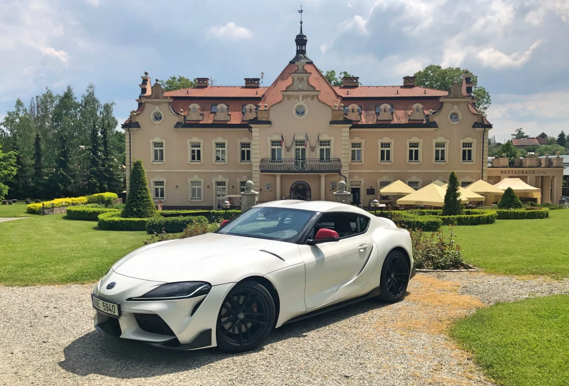Sportovní vůz Toyota GR Supra 2.0 Pure Automatic z roku 2019 v perleťově bílém laku s černými akcenty a vícepaprskovými koly z hliníkové slitiny, zaparkovaný na štěrkové ploše s velkolepou budovou v pozadí. Obrázek zachycuje auto z nízkého předního tříčtvrtečního úhlu, což zvýrazňuje jeho ostré LED světlomety, agresivní styl nárazníků, svažující se střešní linii kupé a výrazné blatníky. Prostředí zahrnuje dobře udržované zelené plochy pod částečně zataženou oblohou, což zvyšuje luxusní a dynamický vzhled scény.