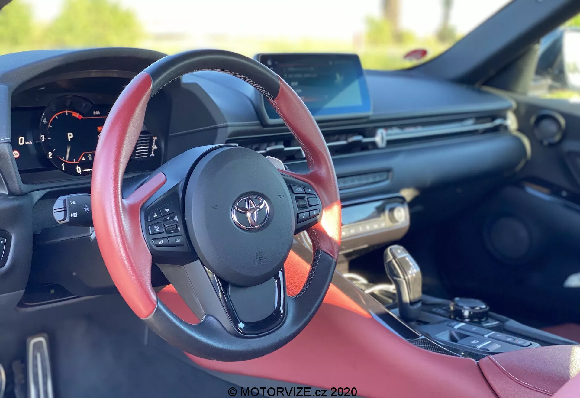 Obrázek je interiérovým záběrem vozu Toyota Supra 2019 (Toyota-GR-Supra-2-0-Pure-Automatic), se zaměřením na kožený volant s červenými akcenty a logem Toyota. Pohled z místa řidiče ukazuje různé ovládací prvky včetně tlačítek pro ovládání audia, hands-free telefonie a tempomatu. Za volantem jsou pádla pro poloautomatickou převodovku. Přístrojový panel zobrazuje otáčkoměr a částečně viditelný digitální displej. Palubní deska obsahuje širokoúhlý informační displej pravděpodobně podporující GPS, přehrávání médií a integraci chytrých telefonů, s větracími otvory a tlačítky pro ovládání klimatizace níže. Středová konzola obsahuje řadicí páku pro automatickou převodovku a volič jízdních režimů pro nastavitelné dynamiky jízdy. Interiérový trim vozu má červené kožené vložky, což naznačuje sportovní nebo luxusní výbavu.