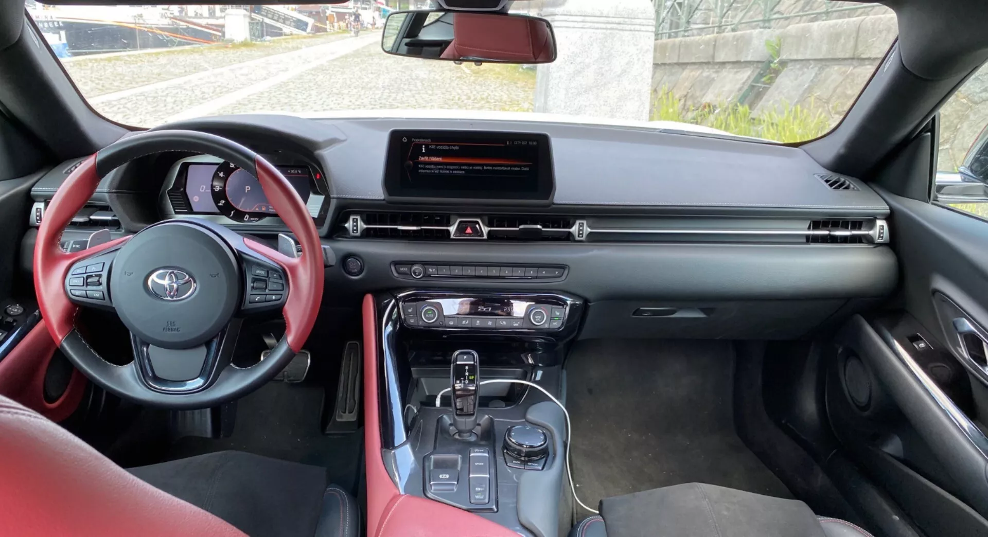 Obrázek zobrazuje interiér moderního vozu Toyota, konkrétně Toyota Supra 2019 (Toyota GR Supra 2.0 Pure Automatic), z perspektivy osoby sedící na sedadle pro zadní pasažéra, mírně směrem k řidiči. Popředí výrazně zobrazuje volant obalený červenou kůží s hmatovými ovladači. Střední panel obsahuje širokoúhlý infotainment displej nahoře, obklopený hmatovými tlačítky a možným digitálním displejem klimatizace. Středová konzole zobrazuje volič převodovky naznačující automatickou převodovku, což možná naznačuje hybridní nebo elektrickou technologii vozidla. Červené okraje předních sedadel naznačují červené čalounění jako estetický prvek této varianty modelu. Denní světlo osvětluje interiér vozu a pohled přes přední čelní sklo ukazuje budovu a oblohu, což potvrzuje, že auto stojí zaparkované.