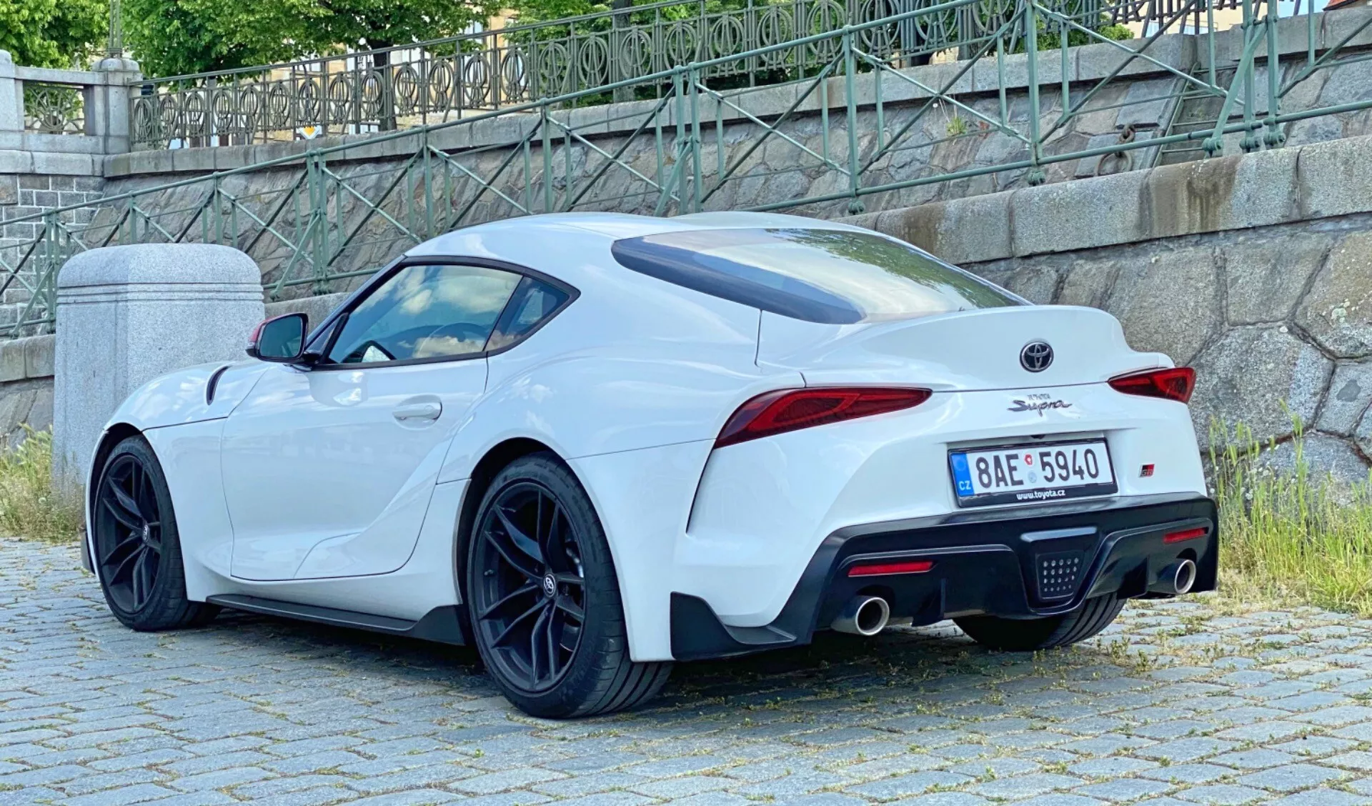 Tři-čtvrtinový zadní pohled na bílý 2019 Toyota GR Supra 2.0 Pure Automatic, představující duální výfukový systém, integrovaný zadní spoiler, LED zadní světla, sportovní vícepaprsková kola, zadní difuzor a boční vzduchové průduchy. Vozidlo je zaparkováno v městském prostředí s kamennou zdí a zelení v pozadí.