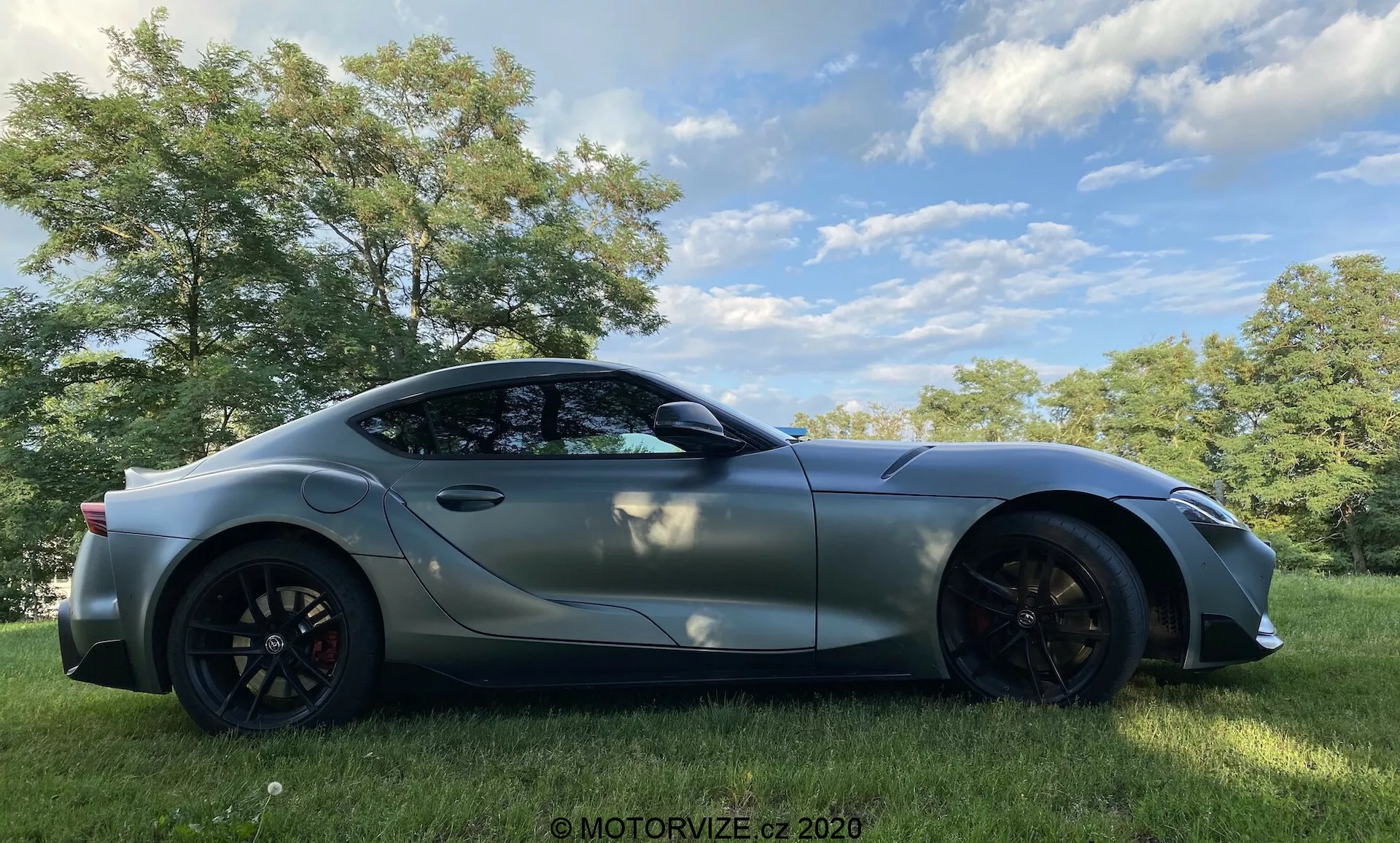 Obrázek sportovního vozu Toyota GR Supra 2.0 Pure Automatic z roku 2019, pořízený z bočního profilu, umístěného venku na travnatém poli s stromy v pozadí, pod polojasnou oblohou. Elegantní, aerodynamický design zahrnuje dlouhou kapotu a sklonenou zadní část, metalický povrch, velká hliníková kola s viditelnými brzdovými třmeny, aerodynamicky tvarovaná boční zrcátka a reflexní tónovaná okna. Zvýrazněné oblouky kol a obrysové čáry podtrhují na výkon orientovaný design tohoto moderního sportovního vozu.