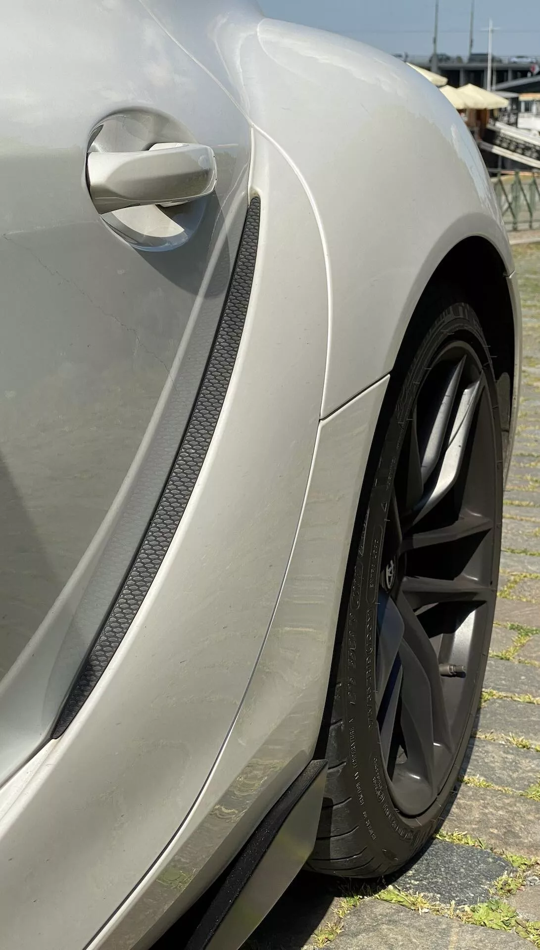 Detailní záběr na přední blatník a oblast kola vozu Toyota GR Supra 2.0 Pure Automatic z roku 2019. Stříbrný metalický lak a elegantní boční zpětné zrcátko auta jsou částečně viditelné. Ventil s uhlíkovým vzorem naznačuje design zaměřený na výkon. Velké černé mnohopaprskové kolo a nízkoprofilová výkonová pneumatika zdůrazňují sportovní vzhled, zatímco odrazný nátěr na blatníku podtrhuje řemeslné zpracování a prvotřídní povrchovou úpravu vozu.