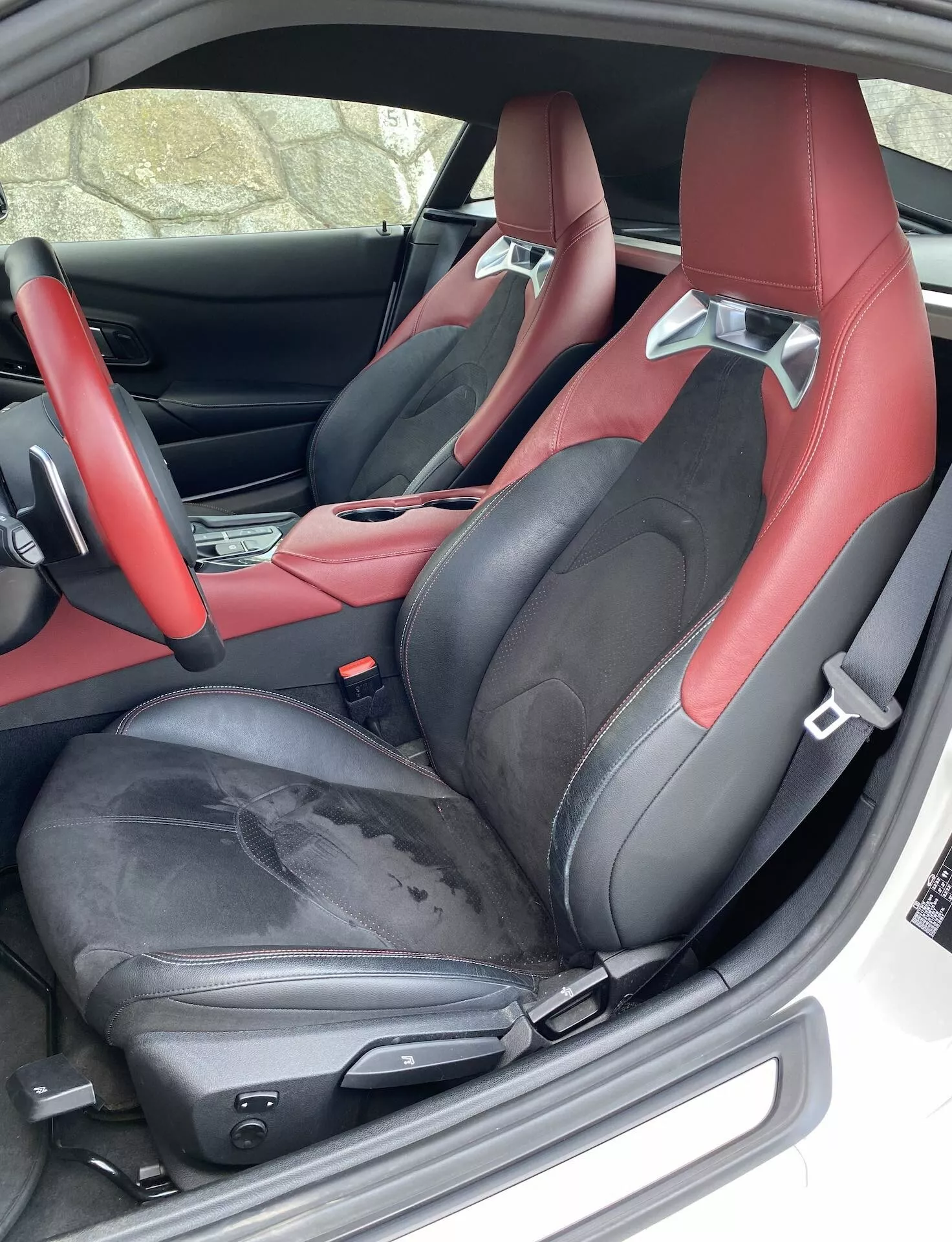 Detailní pohled na interiér z místa řidiče v Toyota GR Supra 2.0 Pure Automatic 2019, ukazující sportovní sedačky s výrazným černo-červeným čalouněním. Sedačky mají integrované opěrky hlavy a boční opěry pro podporu, patrné opotřebení na okraji řidičovy sedačky a elektrické nastavování sedadel, což naznačuje luxus. Viditelná je také část dveří a středové konzoly s odpovídajícím barevným schématem, spolu s výraznou podlahovou rohoží a bezpečnostními pásy.