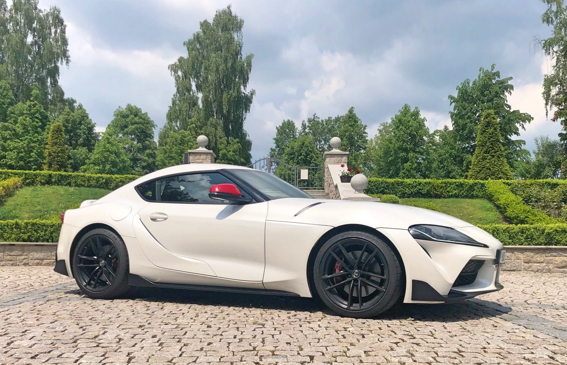 Boční profilový obrázek sportovního auta Toyota GR Supra 2.0 Pure Automatic z roku 2019. Vozidlo stojí na dlážděném povrchu s zelení a upraveným živým plotem v pozadí, což naznačuje park nebo sídlo. Celá délka auta je viditelná z pasažérské strany, což ukazuje jeho elegantní, aerodynamický design s dlouhou kapotou a krátkým zadním krytem. Světle, možná perleťová nebo matně bílá barva auta kontrastuje s černými bočními zrcátky, vstupy vzduchu, koly a zadním difuzorem, což mu dodává agresivní, ale vytříbený vzhled. LED světlomety jsou ostré a úhlové, což odpovídá modernímu vzhledu. Červené brzdové třmeny za víceloučkovými koly naznačují brzdový systém vysokého výkonu. Červený interiér naznačuje luxus, zatímco střešní linie klesá k malému lip spojleru pro aerodynamickou stabilitu.