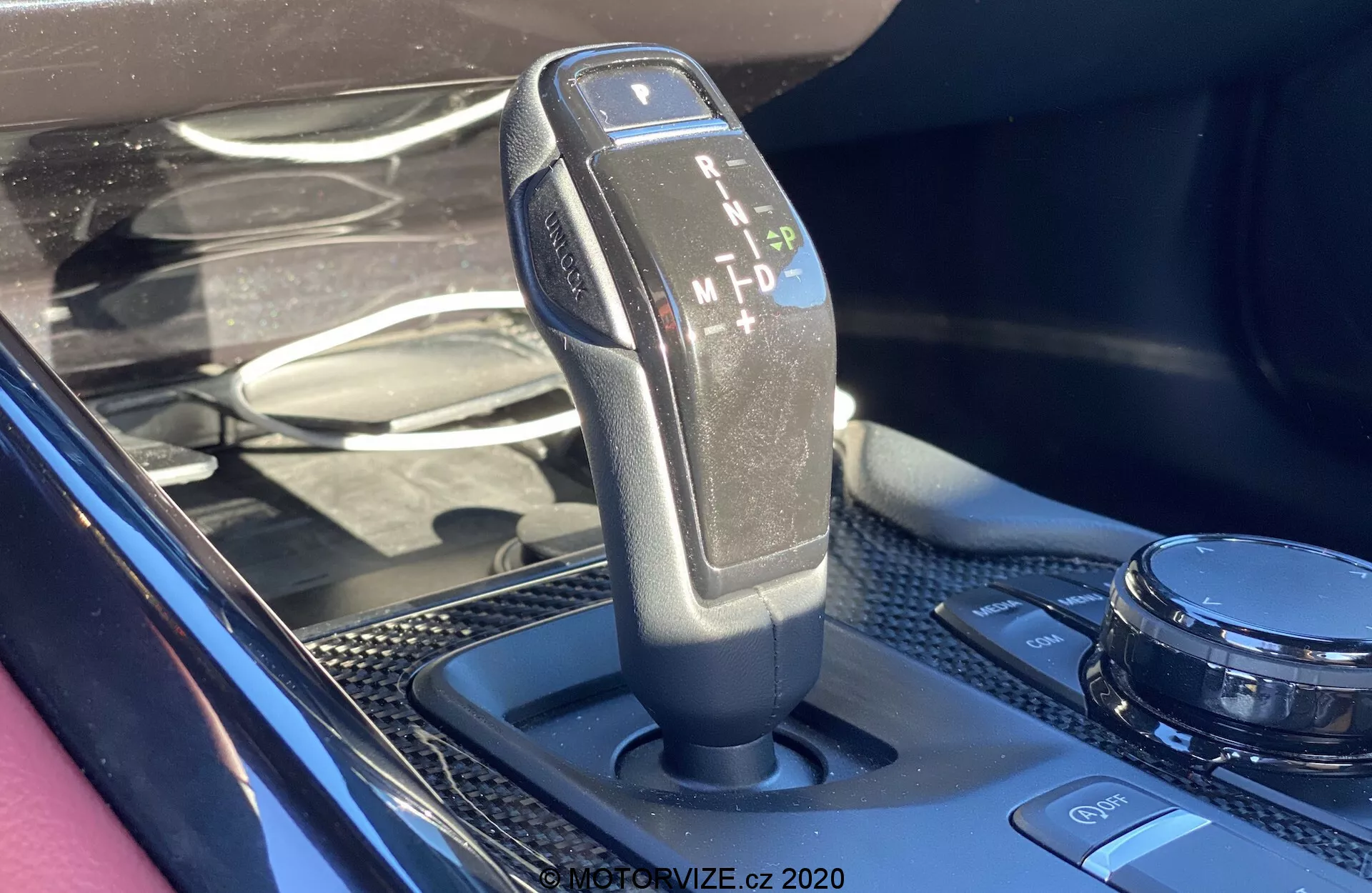 Detailní fotografie automatické řadicí páky vozu Toyota GR Supra 2.0 Pure z roku 2019 s možnostmi parkování (P), zpátečky (R), neutrálu (N), jízdy (D) a manualní/sportovní jízdy (M), s černým plastem a textilním úchytem z kůže na moderního designu středové konzoly s karbonovou úpravou a rotačním ovladačem multimédií.