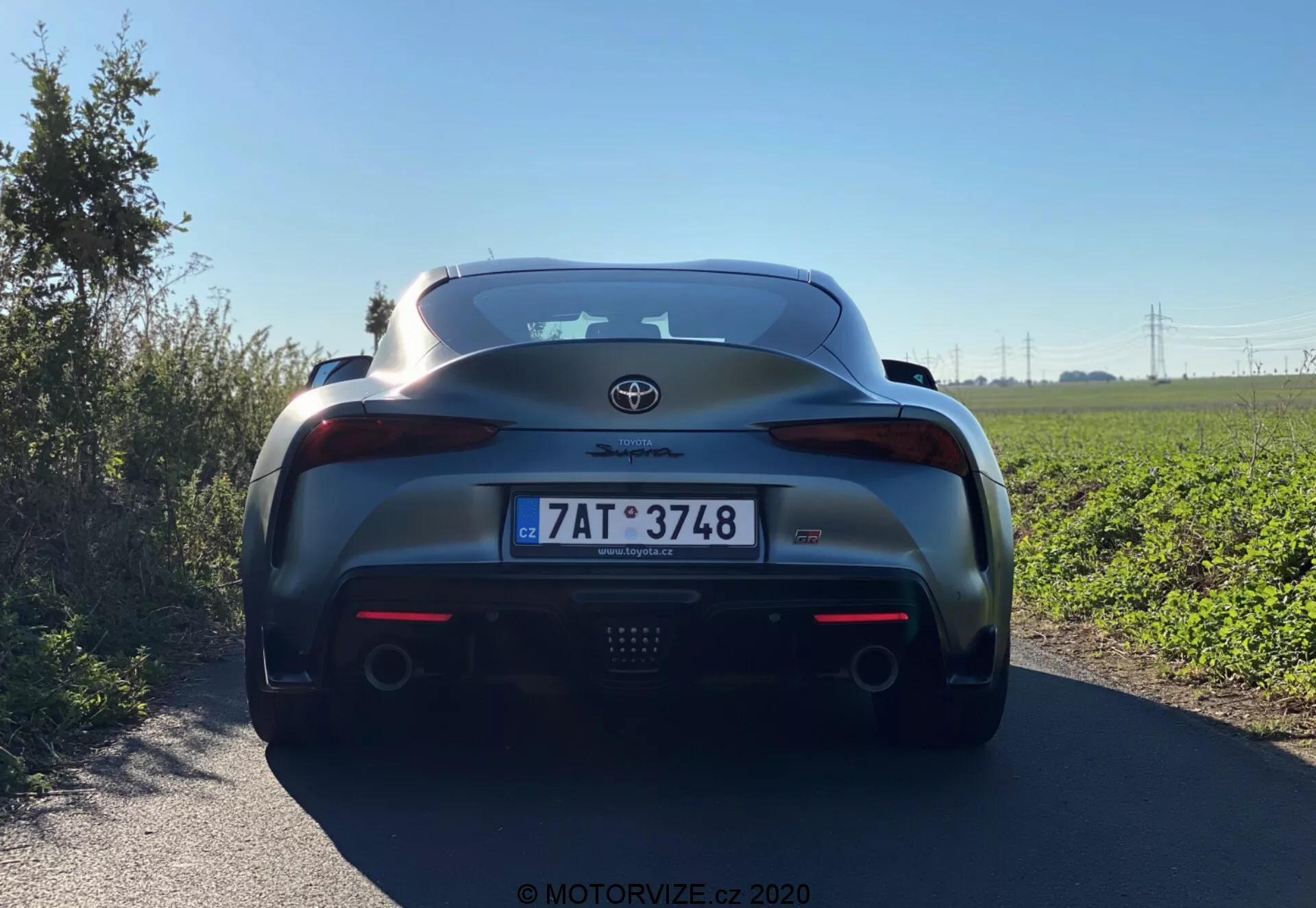 Tento obrázek zobrazuje zadní středový pohled na Toyota GR Supra 2.0 Pure Automatic z roku 2019, zdůrazňující klíčové detaily jako emblém Toyota, nápis Supra, integrovaný zadní spoiler, centrální brzdové světlo, elegantní zadní světelné klustery, dvojité koncovky výfuku, evropskou registrační značku, lesklý metalický lak a černou spodní část zadního nárazníku s prvky podobnými difuzoru.