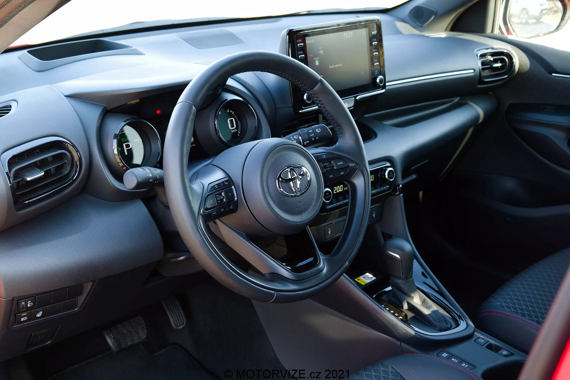 Interiér vozu Toyota Yaris 2020 z pohledu předního spolujezdce se zaměřením na řidičovu oblast. Záběr zahrnuje tříramenný volant s logem Toyota, který disponuje ovládacími prvky pro audio, telefonní rozhraní a nastavení tempomatu. Částečně viditelný je i přístrojový panel vlevo od volantu. Středová konzola obsahuje dotykový infotainment systém s navigací, médiami a nastavením klimatizace. Mezi sedadly je vidět řadicí páka s automatickou převodovkou a tlačítka pro jízdní režimy. Materiály zahrnují kombinaci měkkých plastů a syntetických nebo kožených prvků na klíčových místech dotyku.