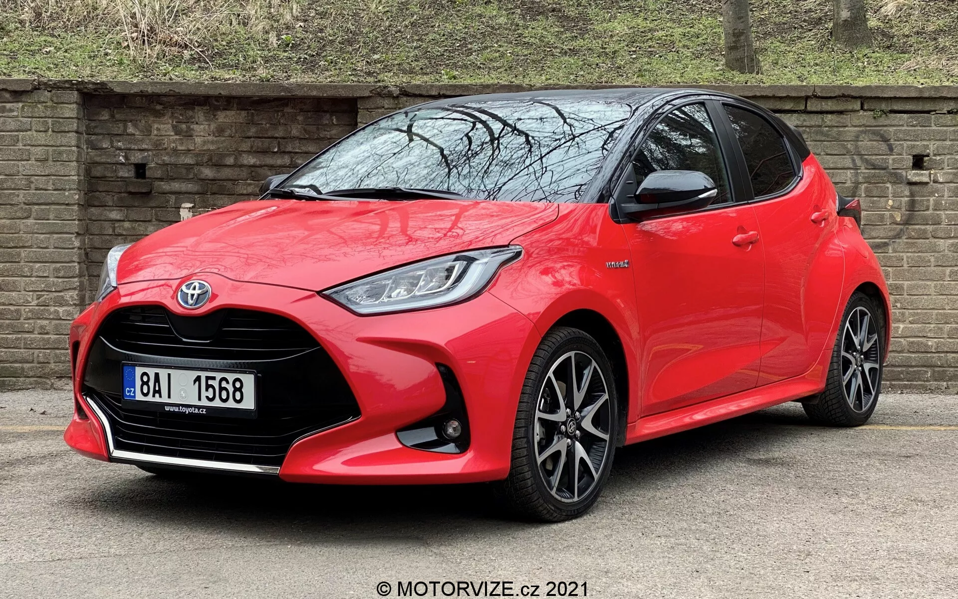 Obrázek ukazuje jasně červenou Toyotu Yaris 2020 zaparkovanou pod úhlem, přičemž čelní levý kvadrant směřuje ke kameře. Pořízený z nízkého úhlu zvýrazňuje elegantní design vozu, včetně LED světlometů směřujících k černě vzorované střední masce chladiče s výrazným emblémem Toyota. Sportovní přední nárazník obsahuje širokou spodní mřížku a černé akcenty. Charakteristické linie podél dveří, integrované směrové signály ve zpětných zrcátkách, černě lemovaná okna a vícesvětná litá kola zdůrazňují moderní, dynamický vzhled auta. Hybridní odznak označuje pokročilou ekologickou technologii.