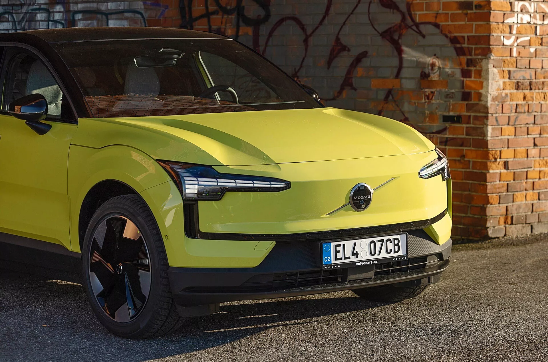 Limo zelený elektrický vůz Volvo EX30 rok 2024 zaparkovaný před zdí s graffiti, ukazuje aerodynamický design, moderní LED světlomety a absenci přední mřížky