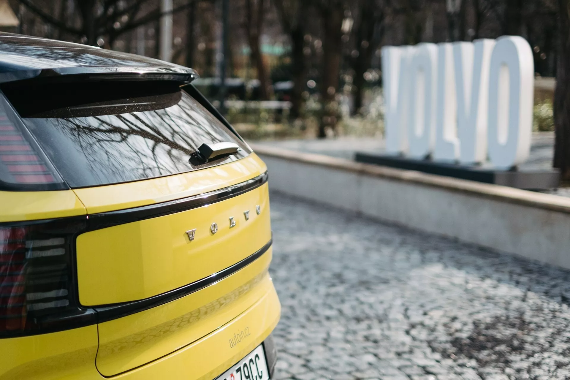 Detailní pohled na zadní část žlutého Volvo EX30 2024 s horizontálními LED zadními světly, možnou zadní kamerou nebo senzory, jedním stěračem zadního skla, označením EV nebo hybridu a anténou ve tvaru žraločí ploutve, zaparkované na dlažebním povrchu s rozmazaným nápisem 'OLVO' na pozadí