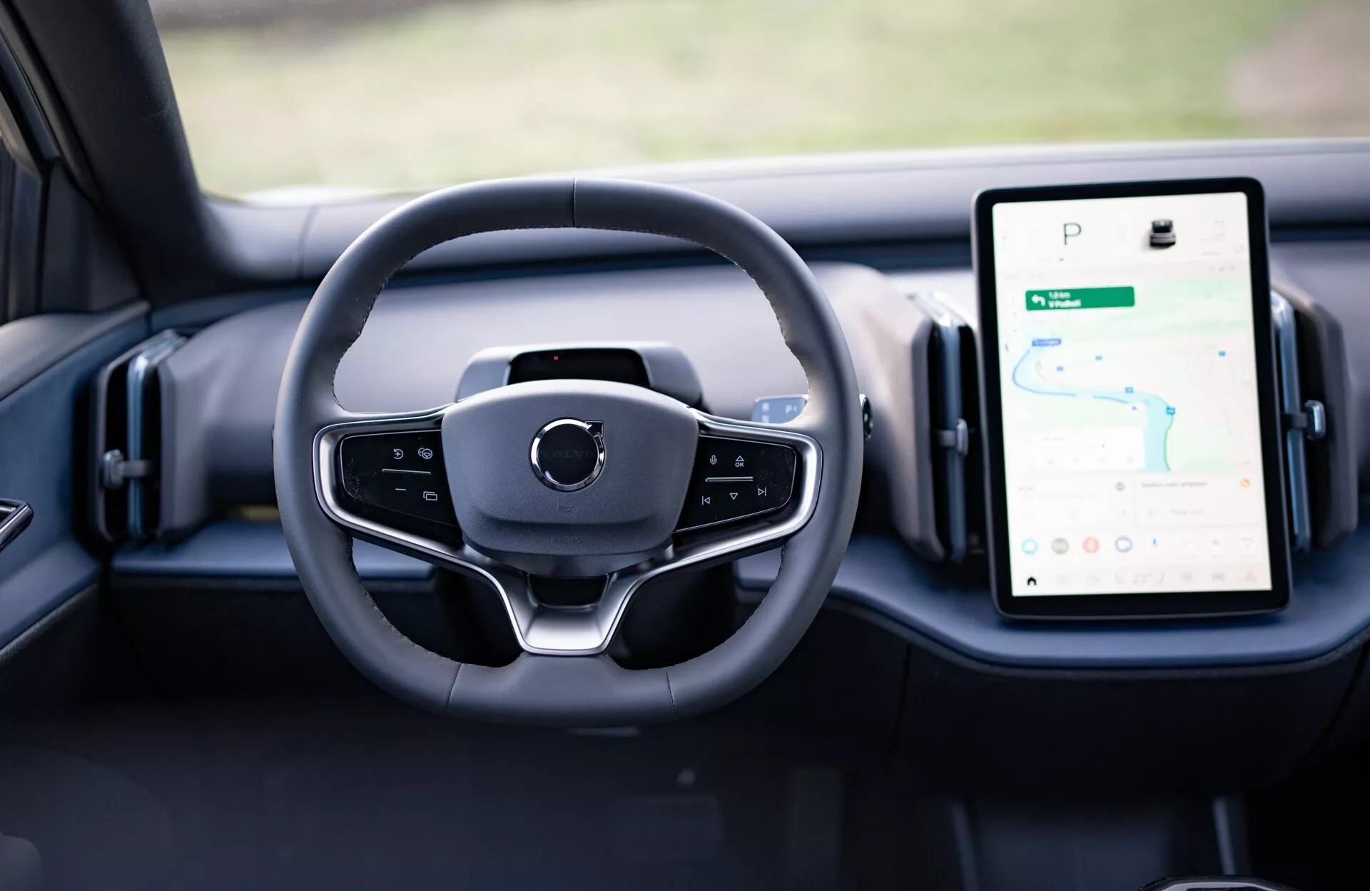 Interiérový pohled na Volvo EX30 2024 ukazující moderní design volantu a digitální ciferníky na palubní desce, včetně vertikálně orientovaného dotykového infotainment systému s GPS.