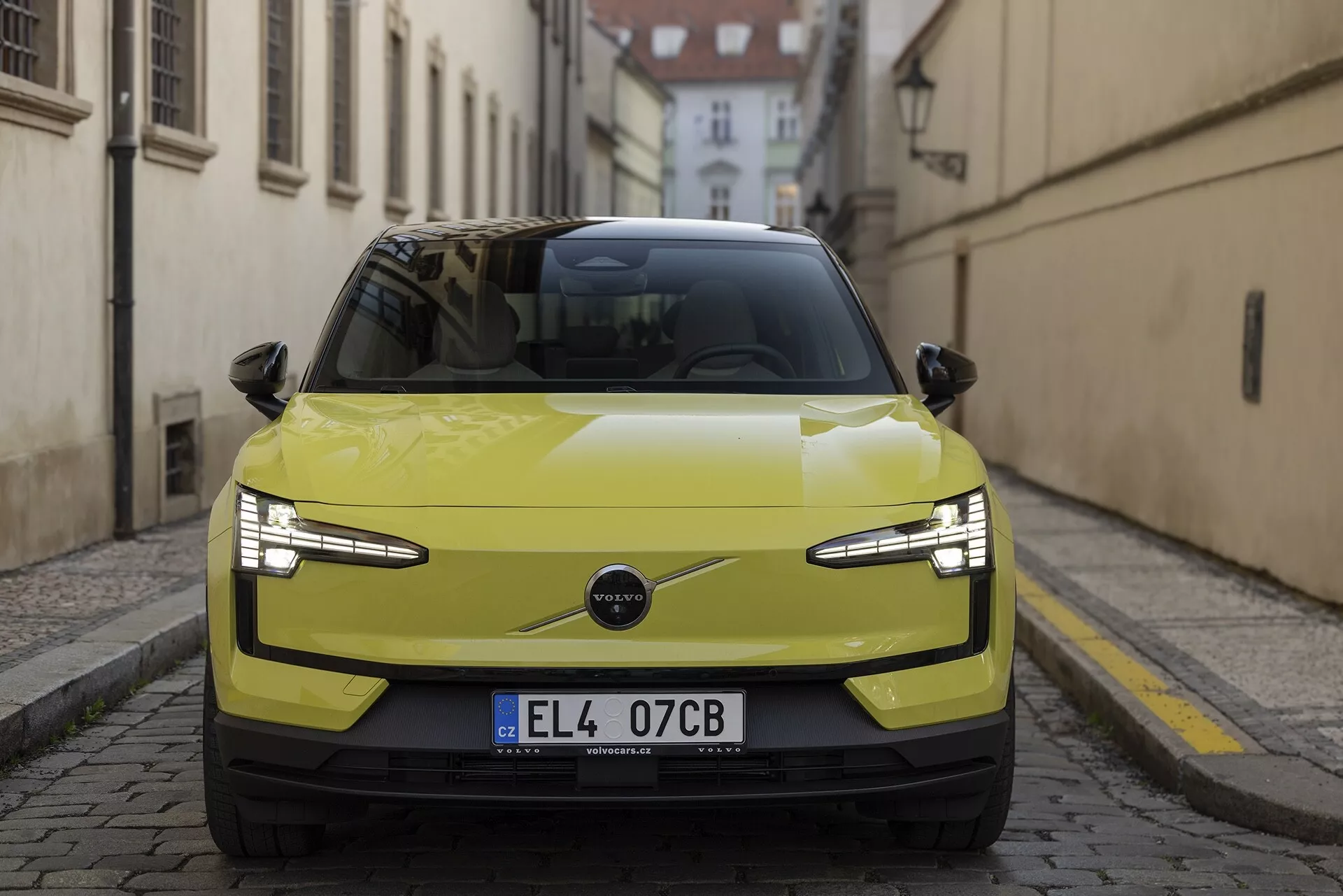 Přední pohled na Volvo EX30 2024 v limetkově žluté barvě, zdůrazňující jeho elegantní LED světlomety, minimalistickou mřížku naznačující elektrický pohon a pokročilé technologické funkce jako parkovací senzory a možná ADAS, na pozadí historické evropské ulice