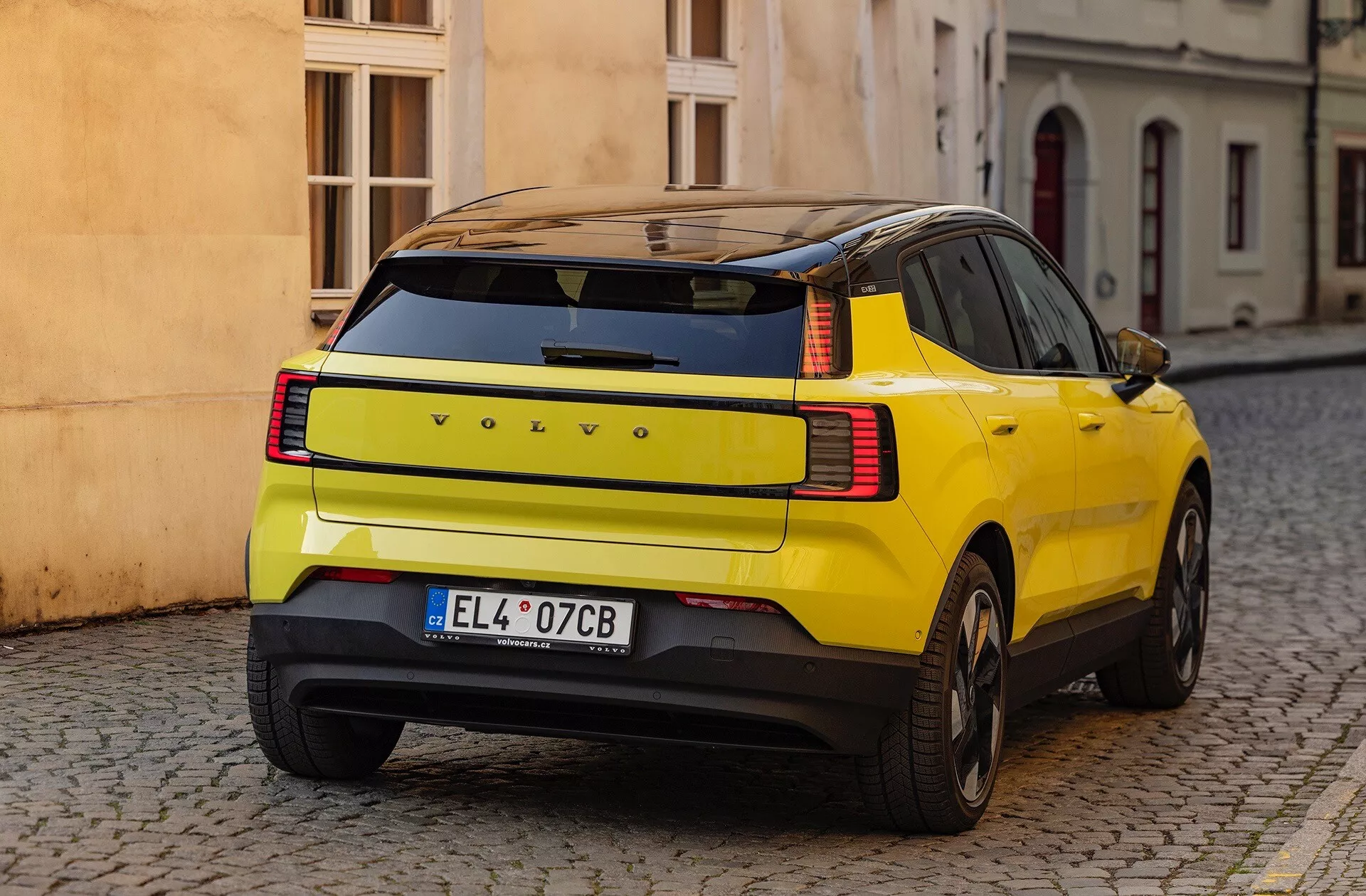 Žluté SUV Volvo EX30 2024 ukazuje své charakteristické zakřivení zadního blatníku, LED zadní světla a bezpečnostní prvky, zaparkováno na dlážděné ulici z pohledu ze tří čtvrtin zezadu