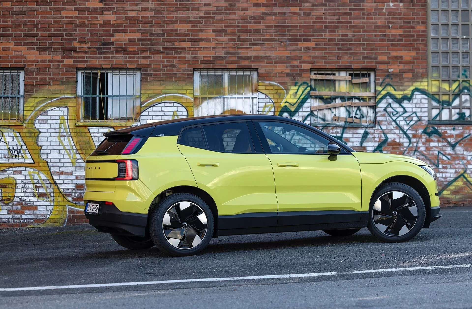 Boční pohled na limetkově zelené SUV Volvo EX30 ročník 2024 zaparkované u obrubníku městské ulice s graffiti uměním na pozadí, předvádějící sportovní černá slitinová kola, LED zadní světla a vysoko umístěnou brzdovou svítilnu.