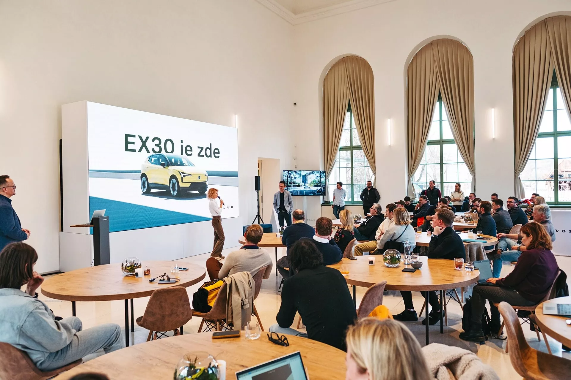Prezentace elektrického vozidla Volvo EX30 2024 ve prostorné místnosti s vysokými stropy a vysokými obloukovými okny, s žlutým EX30 zobrazeným z pohledu tři čtvrtiny zepředu na velké obrazovce