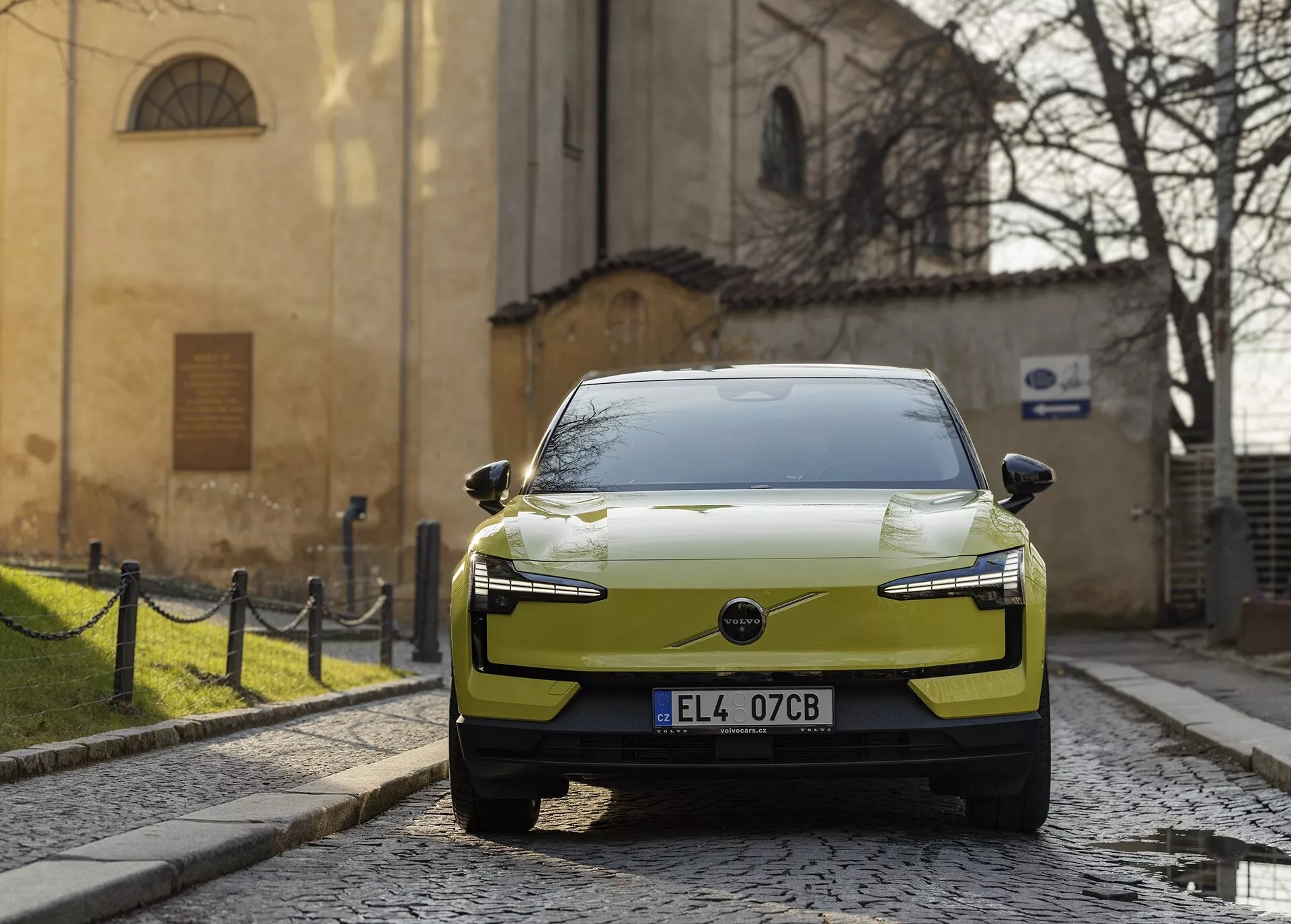 Moderní elektrické auto Volvo EX30 rok 2024 v chartreusové barvě zaparkované na dlažební ulici, zobrazené z pravé přední perspektivy s LED světlomety a aerodynamickým designem kapoty na pozadí historické evropské architektury.