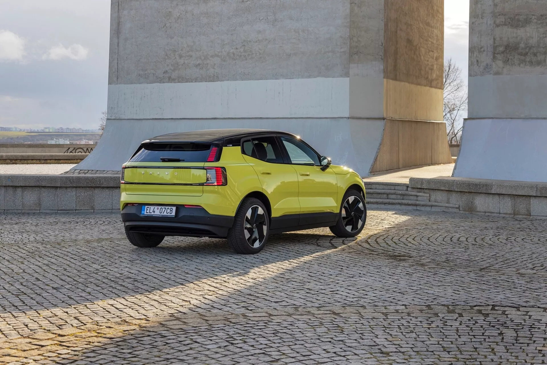 Lime-zelené SUV Volvo EX30 2024 v pohledu ze tří čtvrtin odzadu ukazující boční profil, zadní světla, zavazadlový prostor a černá slitinová kola poblíž kamenných zdí na dlažebním povrchu