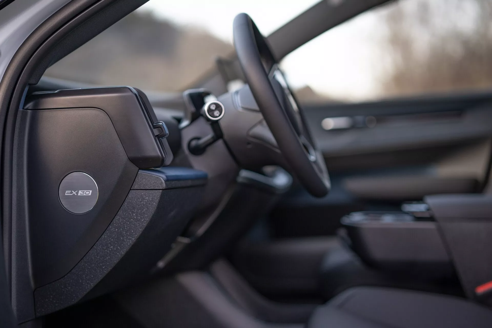 Interiérový pohled na Volvo EX30 2024 zaměřený na stranu řidiče s detailním sportovním nebo prémiovým designem sedadla, audio systém Harman Kardon na sloupku B, multifunkční volant a náznaky pokročilého informačního systému a designu dveřního panelu spolujezdce