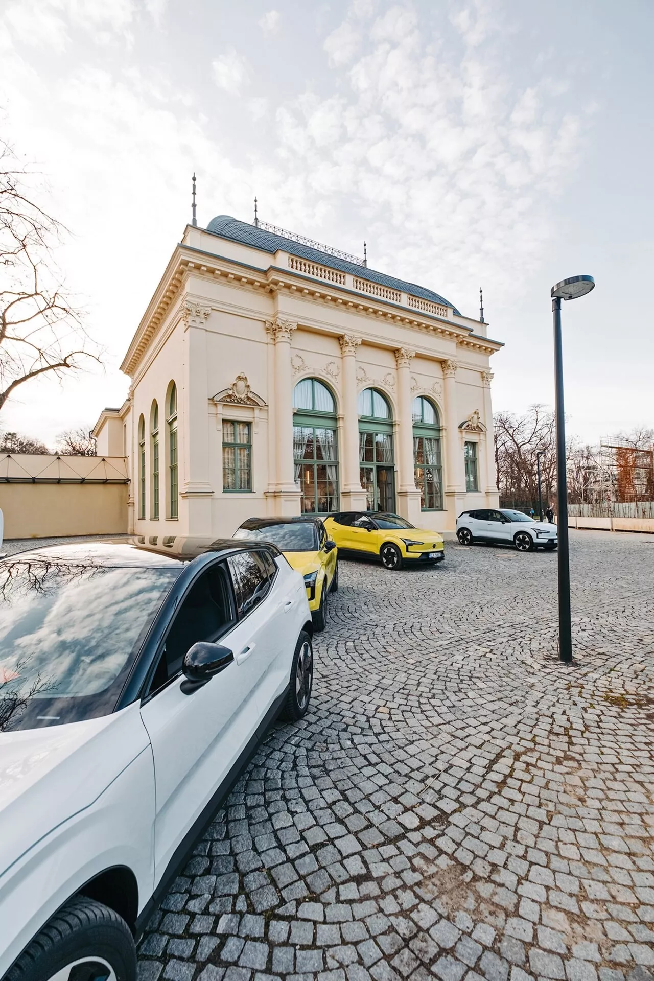 Městská scéna se třemi auty a elegantní budovou na pozadí, pořízená z širokoúhlé perspektivy zvýrazňující bílý současný sedan, výrazné žluté sportovní auto a bílý sedan nebo hatchback ve světle pozdního odpoledne