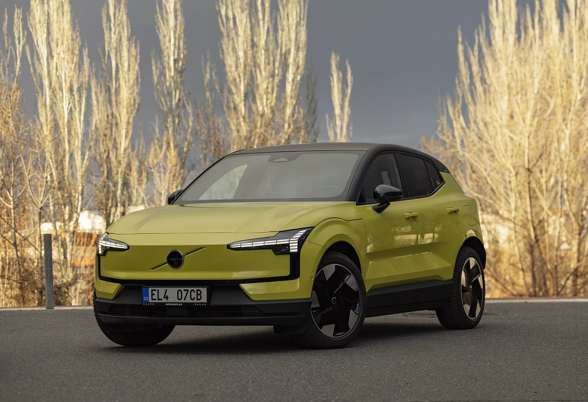 Moderní limetkově zelený elektrický vůz Volvo EX30 2024 ve tříčtvrtečním předním pohledu zdůrazňující jeho elegantní LED světlomety, absenci tradiční mřížky pro lepší aerodynamiku, velká černá slitinová kola, karoserií lakovaná vnější zrcátka s potenciální integrovanou technologií, hladký a aerodynamický boční profil s vzhledem podobným kupé a živý metalický povrch, zaparkovaný v přírodním prostředí