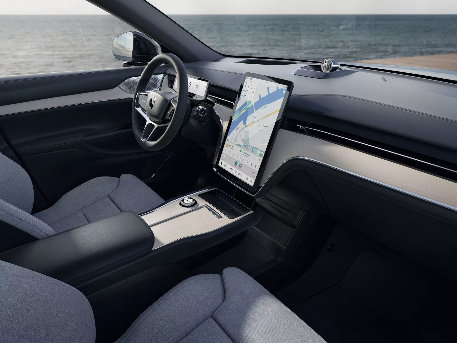 Interiérový pohled na Volvo EX90 2023 ukazující oblast řidiče a středovou konzolu s elevovaným pohledem, obsahující multifunkční volant obalený kůží, velký vertikálně orientovaný dotykový informační systém s GPS, větrací otvory klimatizace, akcenty z kartáčovaného kovu, sedadla potažená látkou a elegantní úložné prostory.
