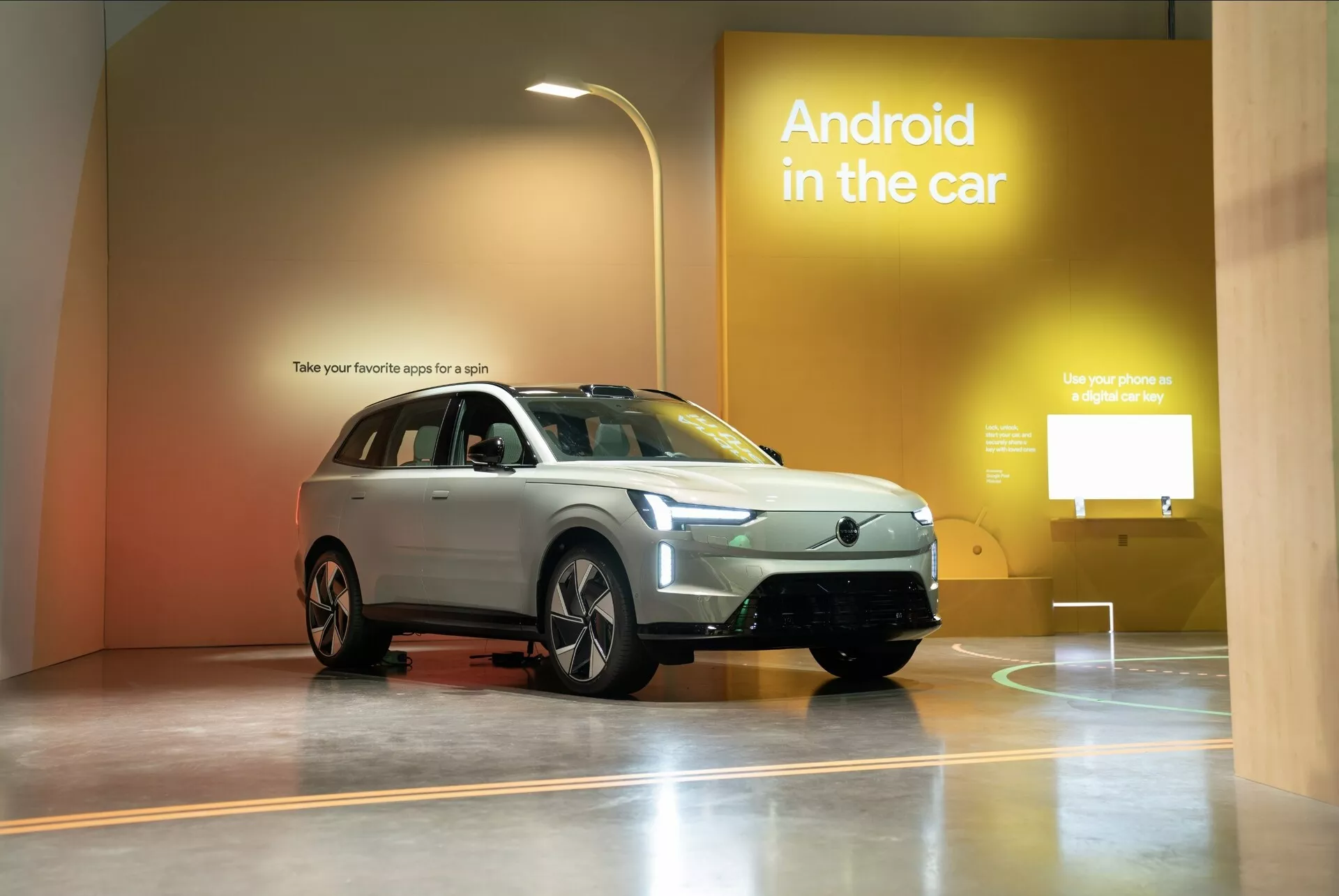 Volvo EX90 první generace roku 2023 vystaveno v showroomu, zdůrazňující jeho SUV styl, pohled z přední části vozidla ukazující mřížku, světlomety, přední nárazník, kapotu, kola, boční zrcátka a okna s reklamou na technologii Android v autě na pozadí