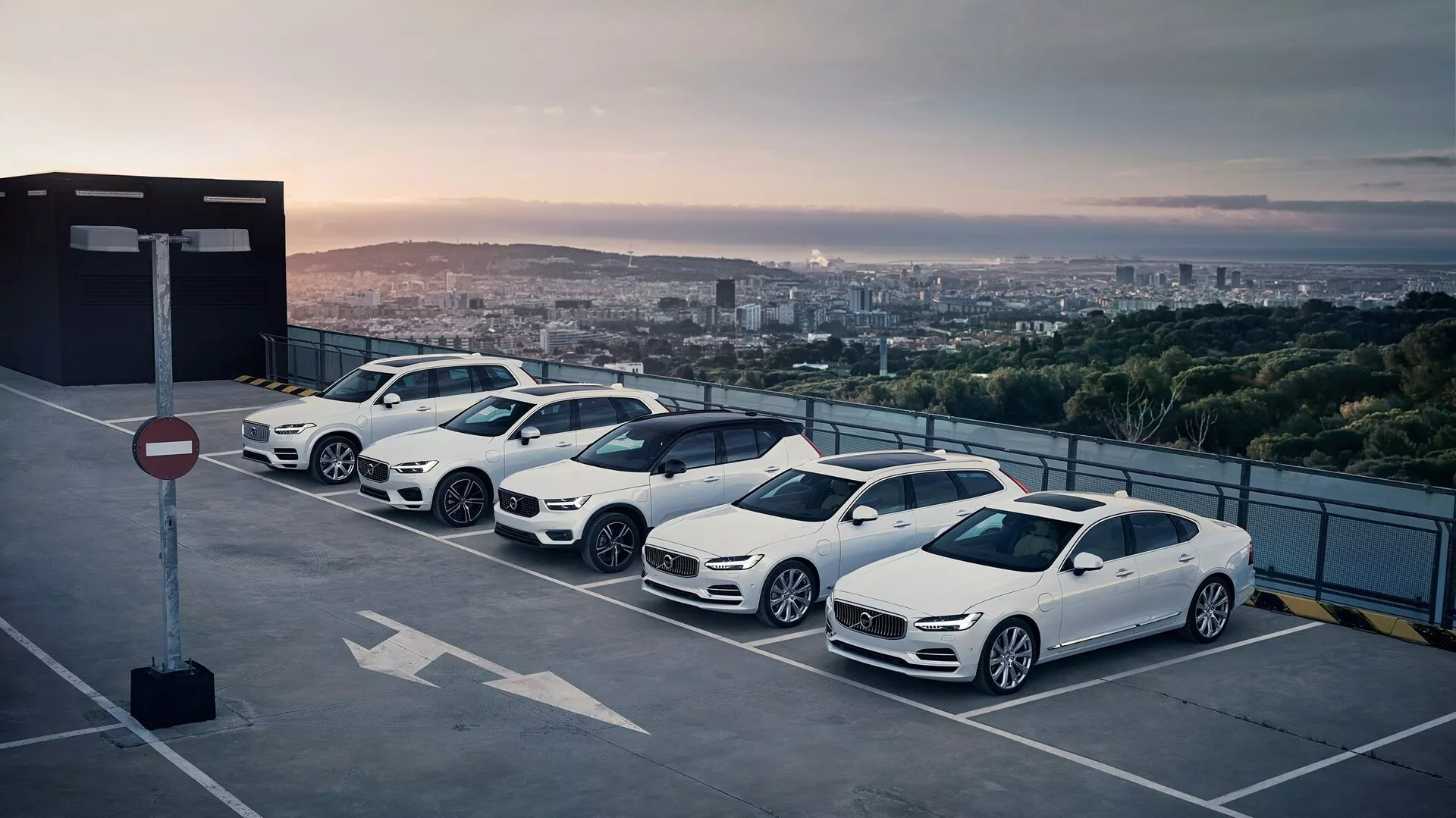 Pohled z výšky na čtyři vozy Volvo XC90 2015 (2. generace) zaparkované diagonálně na střeše během východu nebo západu slunce, zvýrazňující jejich pravé strany s panoramatickým výhledem na město.